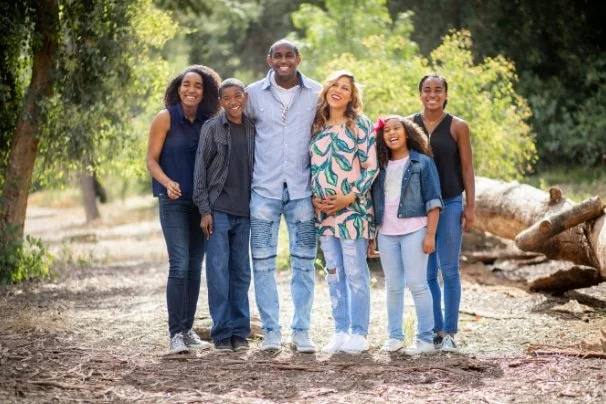 A smiling family of six stands together outdoors in a wooded area, posing for a photo on a sunny day.