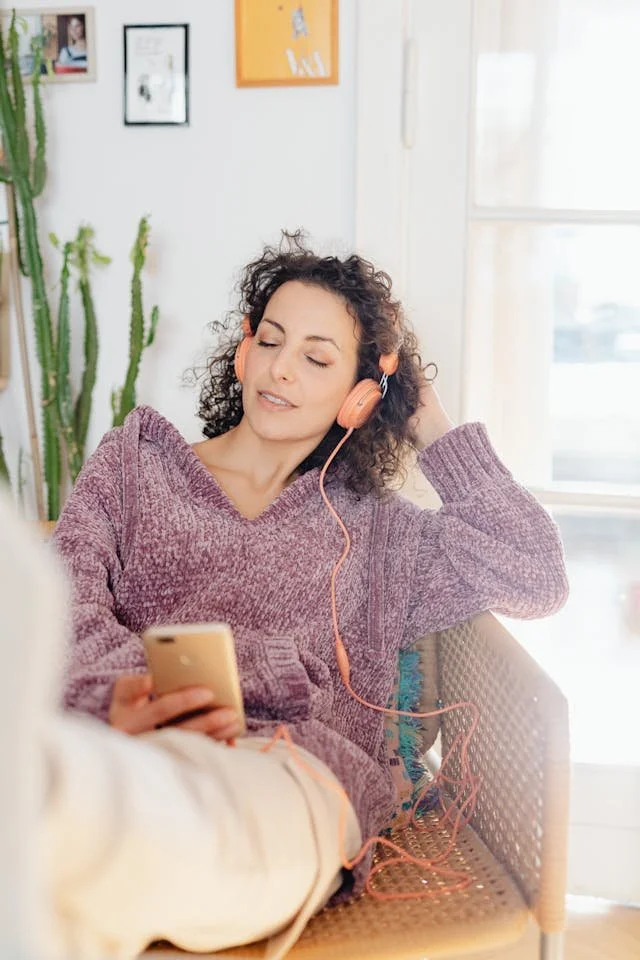 Person in a purple sweater relaxing on a chair, listening to music with earphones and holding a smartphone in a bright, cozy room.