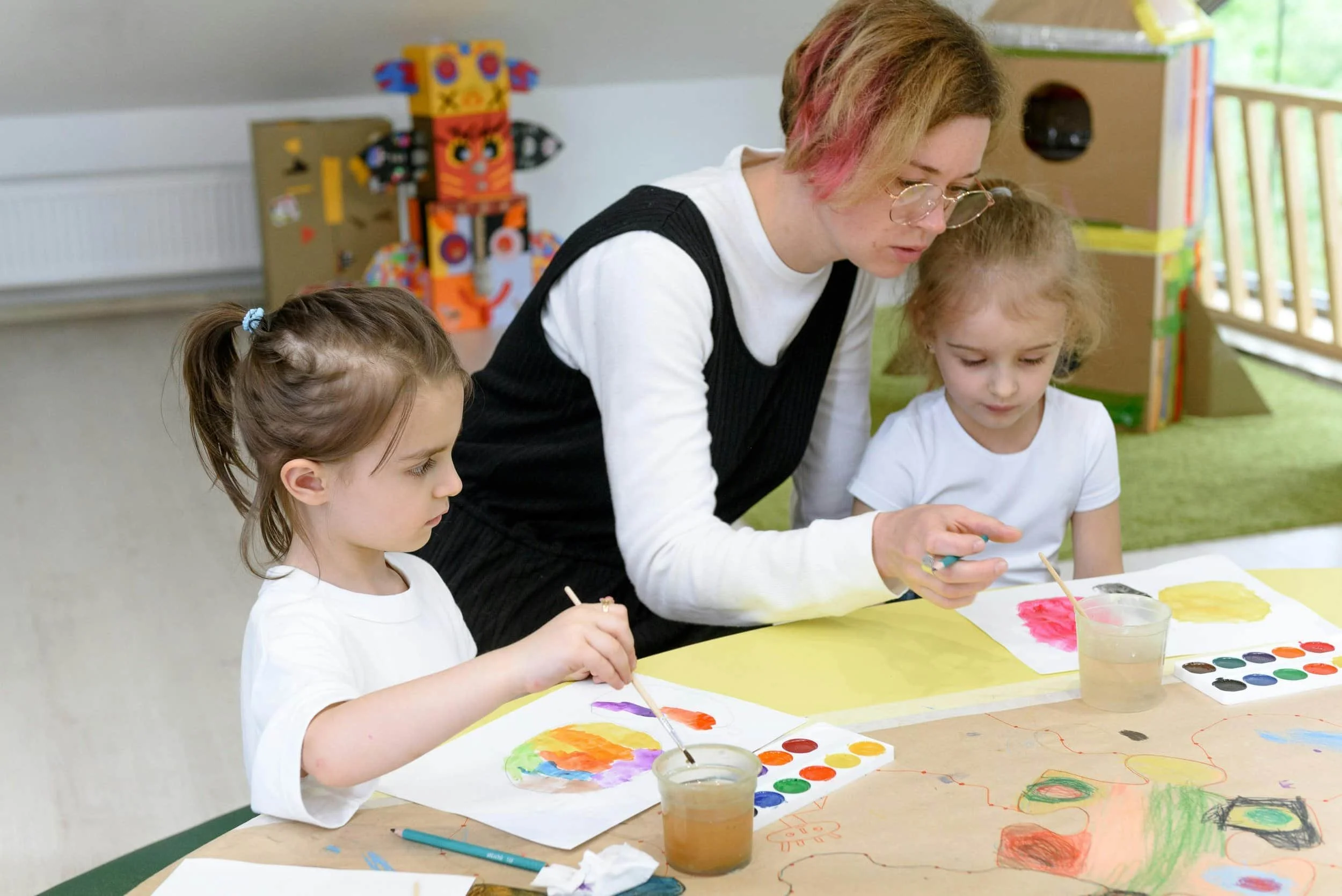 Adult and two children painting with watercolors at a table in a brightly lit classroom with colorful crafts in the background.