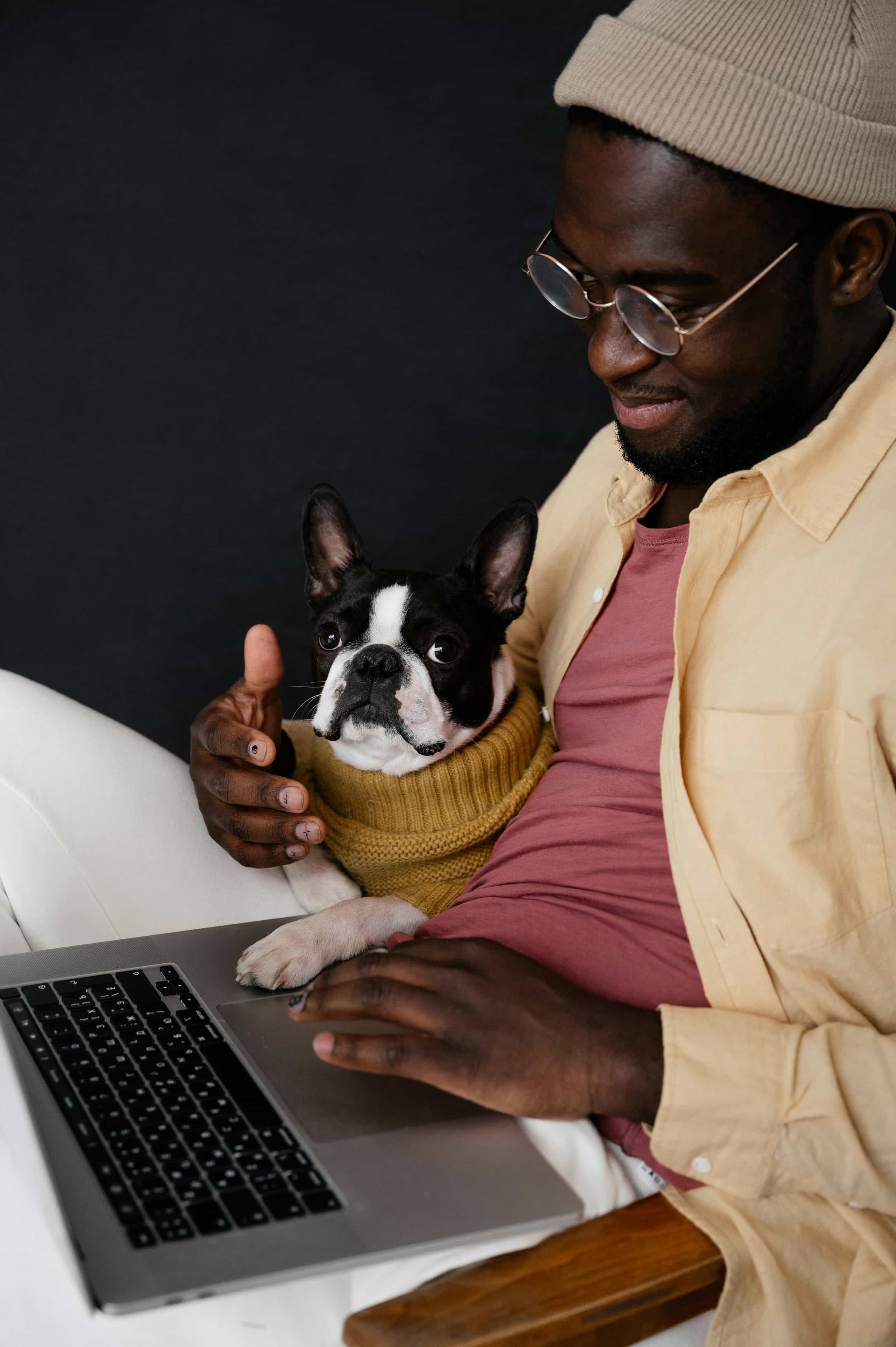 A person sits comfortably with a small dog in a cozy yellow sweater, typing on a laptop, against a dark background.