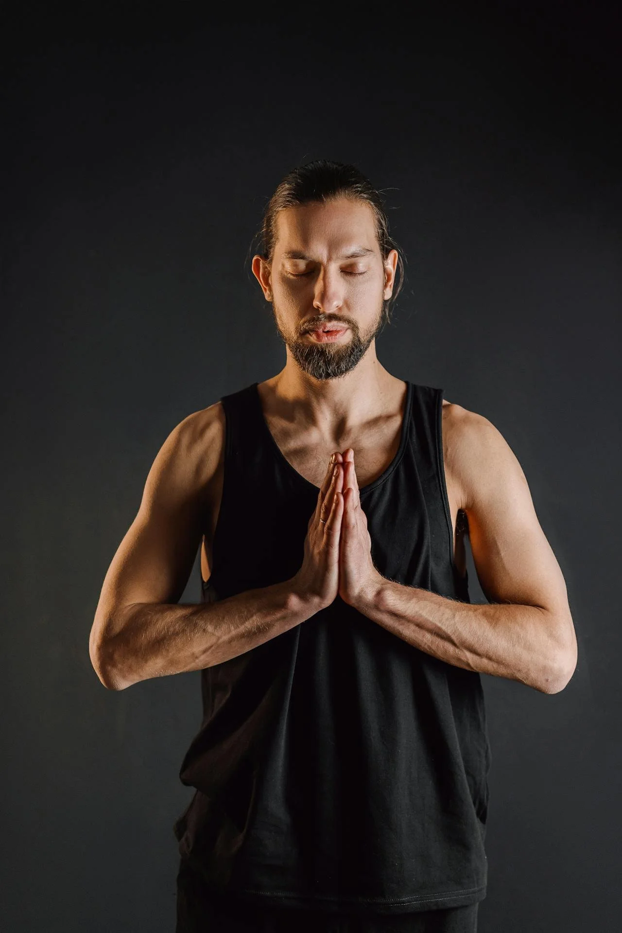 Man in black tank top with hands pressed together at chest, posing in front of a dark background.