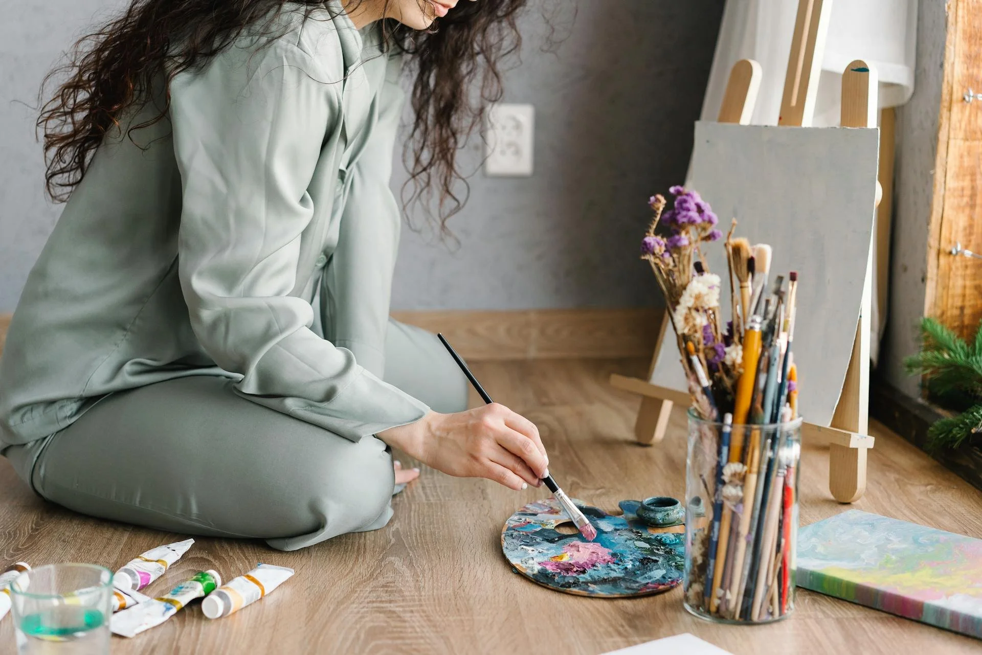 Person in green clothes kneeling on wooden floor, mixing paint on palette near blank canvas and jar of brushes.