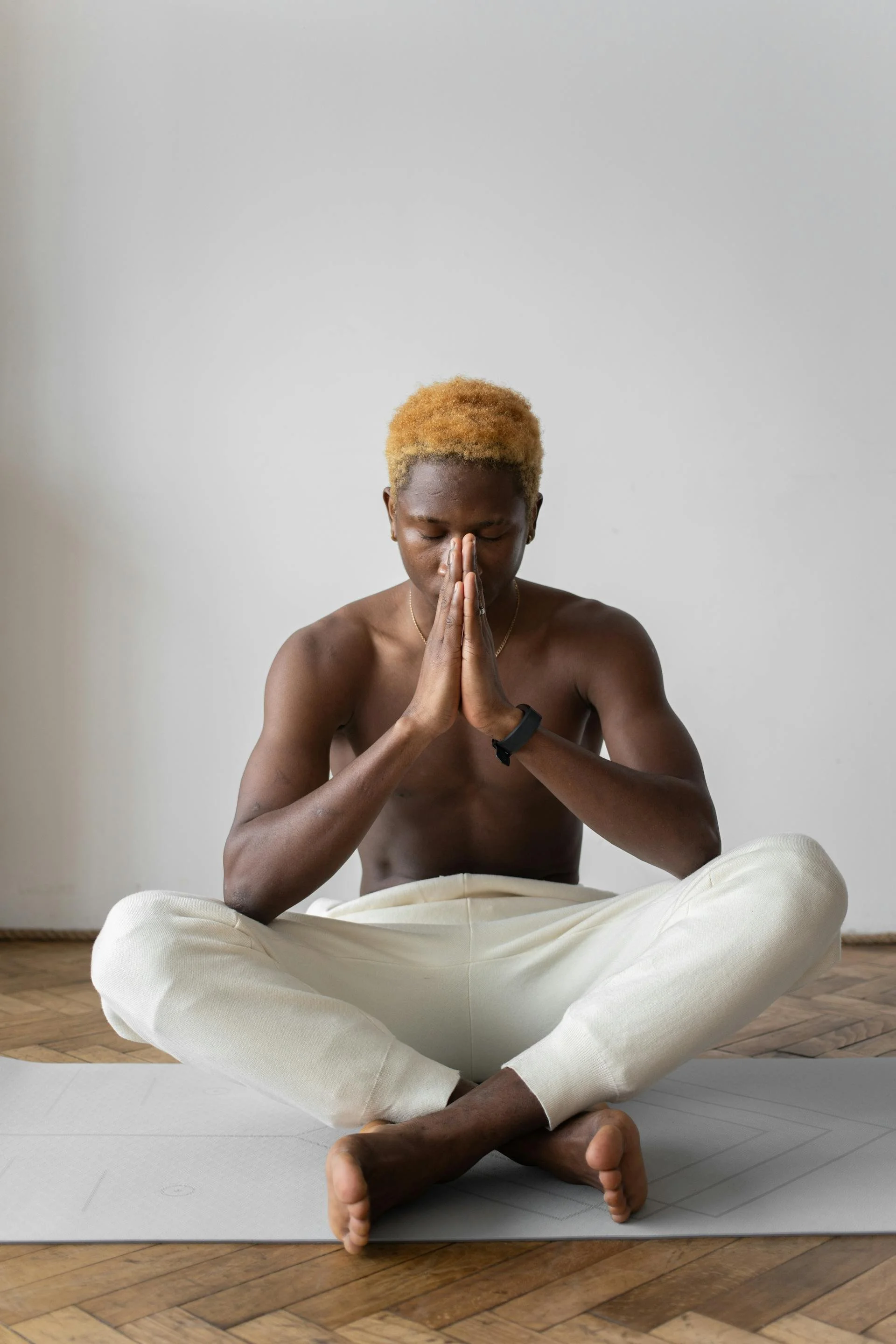 A man in white pants sits crosslegged with his eyes closed and hands clasped together in front of him in namaste
