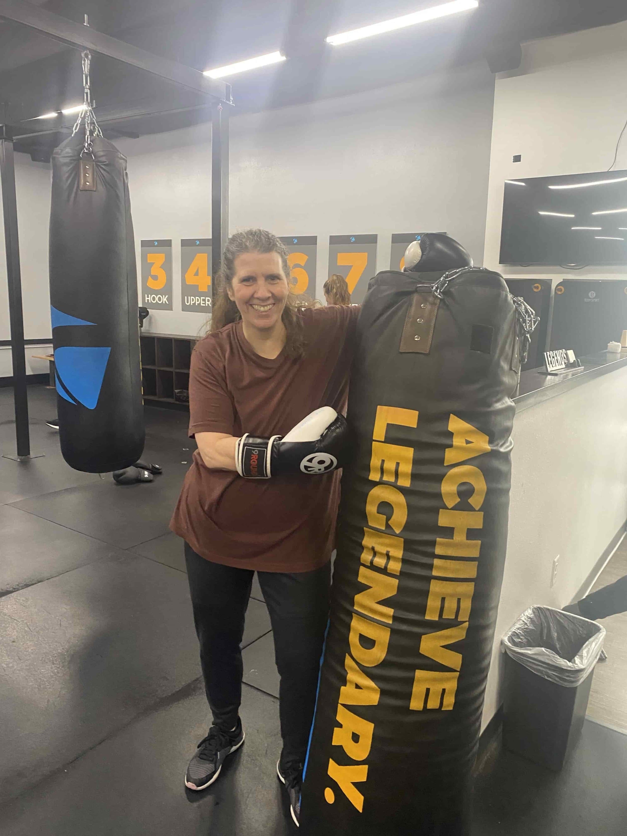 A person in boxing gloves leans on a heavy bag labeled "ACHIEVE LEGENDARY" in a gym, with training equipment in the background.