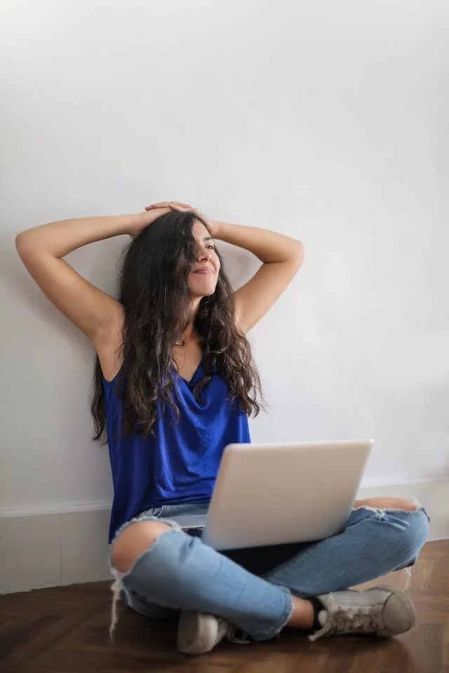 Woman in a blue top and ripped jeans sits on the floor with a laptop, smiling and resting her hands on her head.