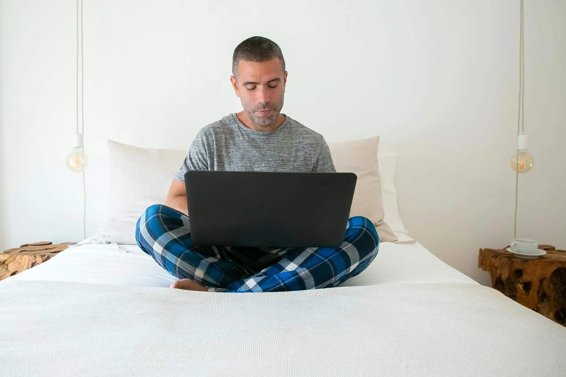 Person sitting cross-legged on a bed, wearing pajamas and using a laptop, with pendant lights and wooden side tables in the background.