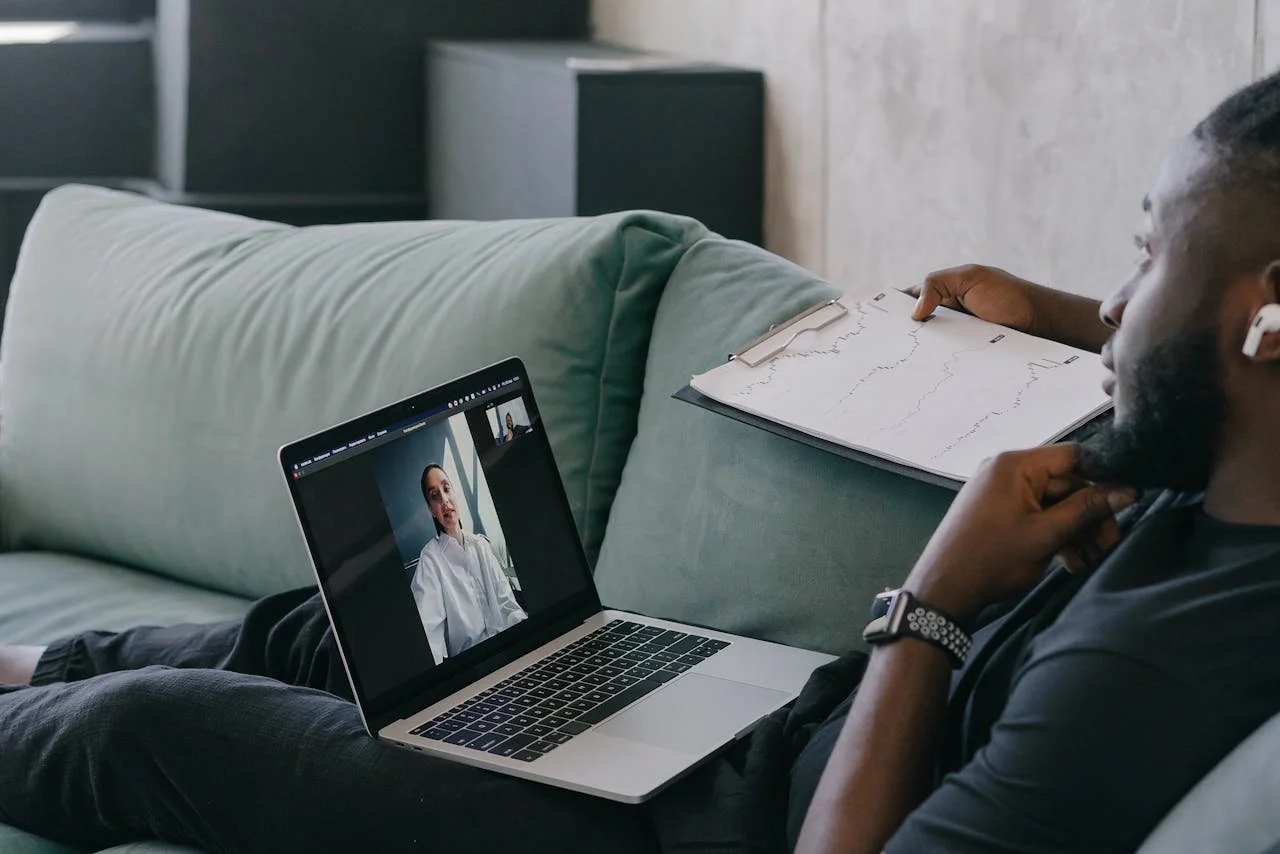 Person sitting on a green couch, video calling on a laptop while holding a clipboard with graphs or charts.