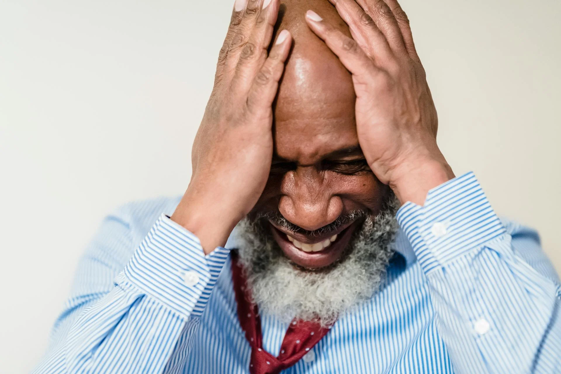 Older man with a gray beard wearing a blue striped shirt and red tie, holding his head in frustration or stress.
