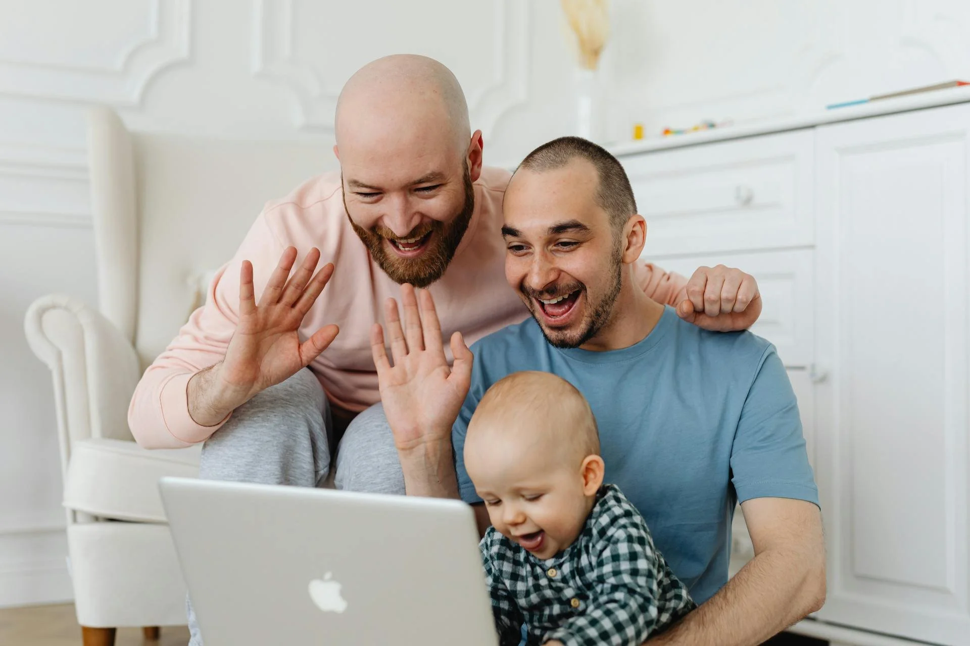Two men and a baby sitting by a laptop, smiling and waving during a video call in a bright living room.