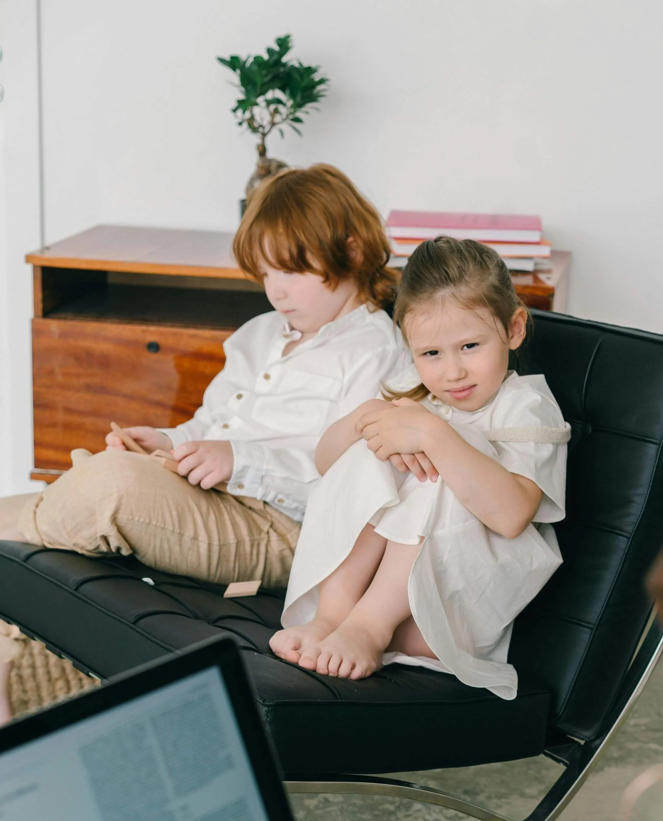 Two children in casual white outfits sit closely together on a black chair, surrounded by a modern, minimalistic room.