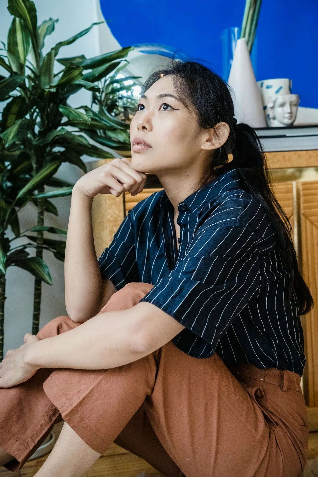 Person sitting on the floor, wearing a navy blue striped shirt and rust-colored pants, next to a green plant and wooden cabinet.