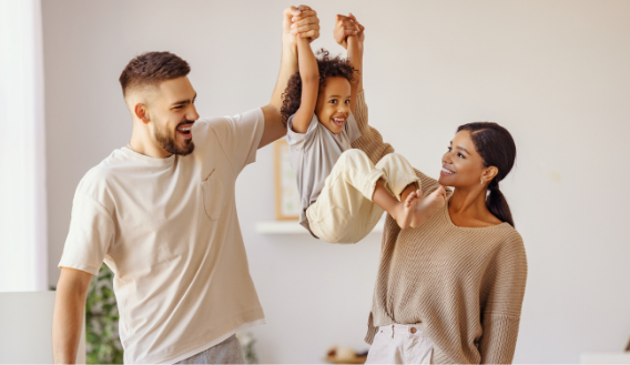 Parents joyfully lift their child by the hands in a bright, cozy living room.