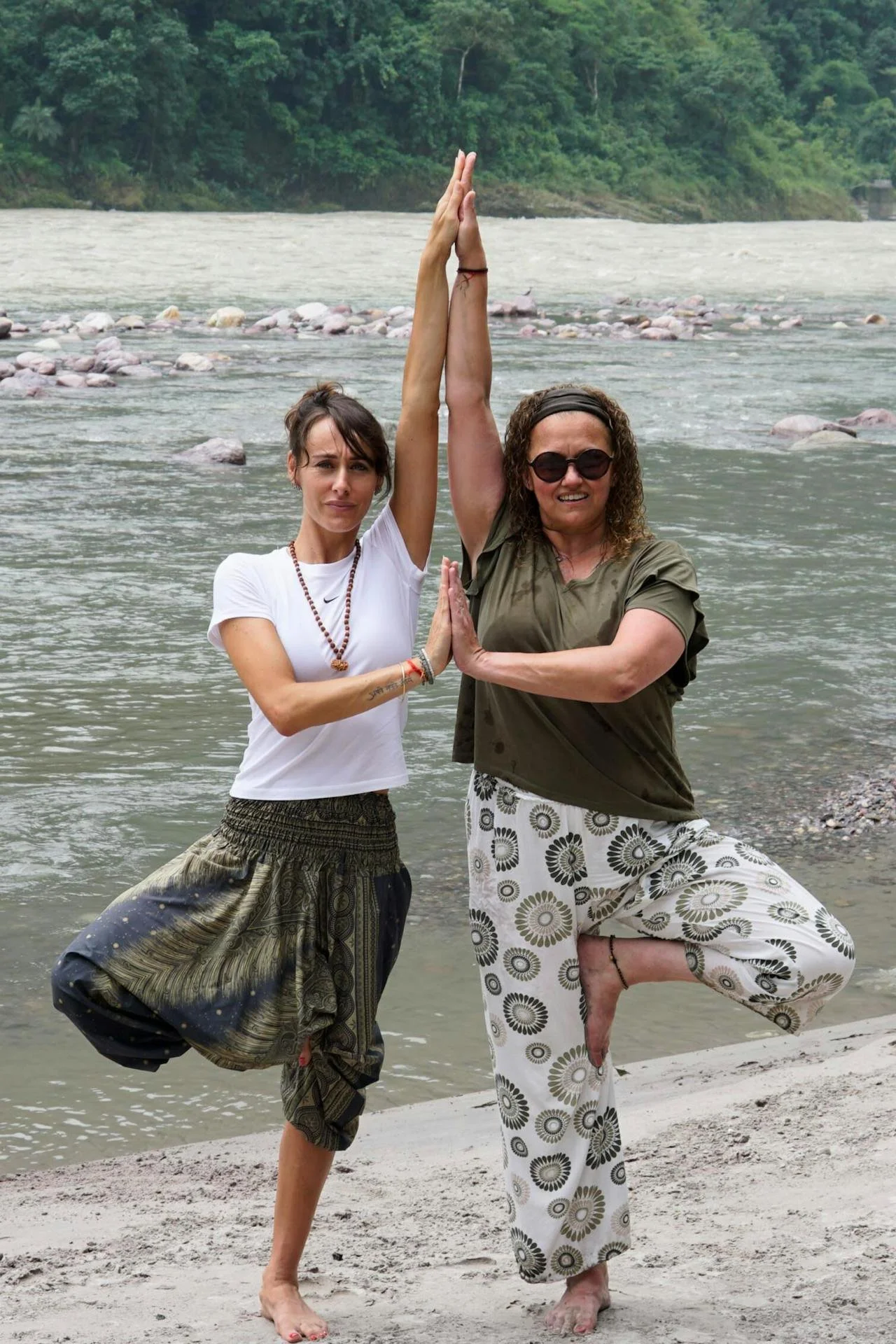 Two women stand together in a yoga tree pose on the side of a beach