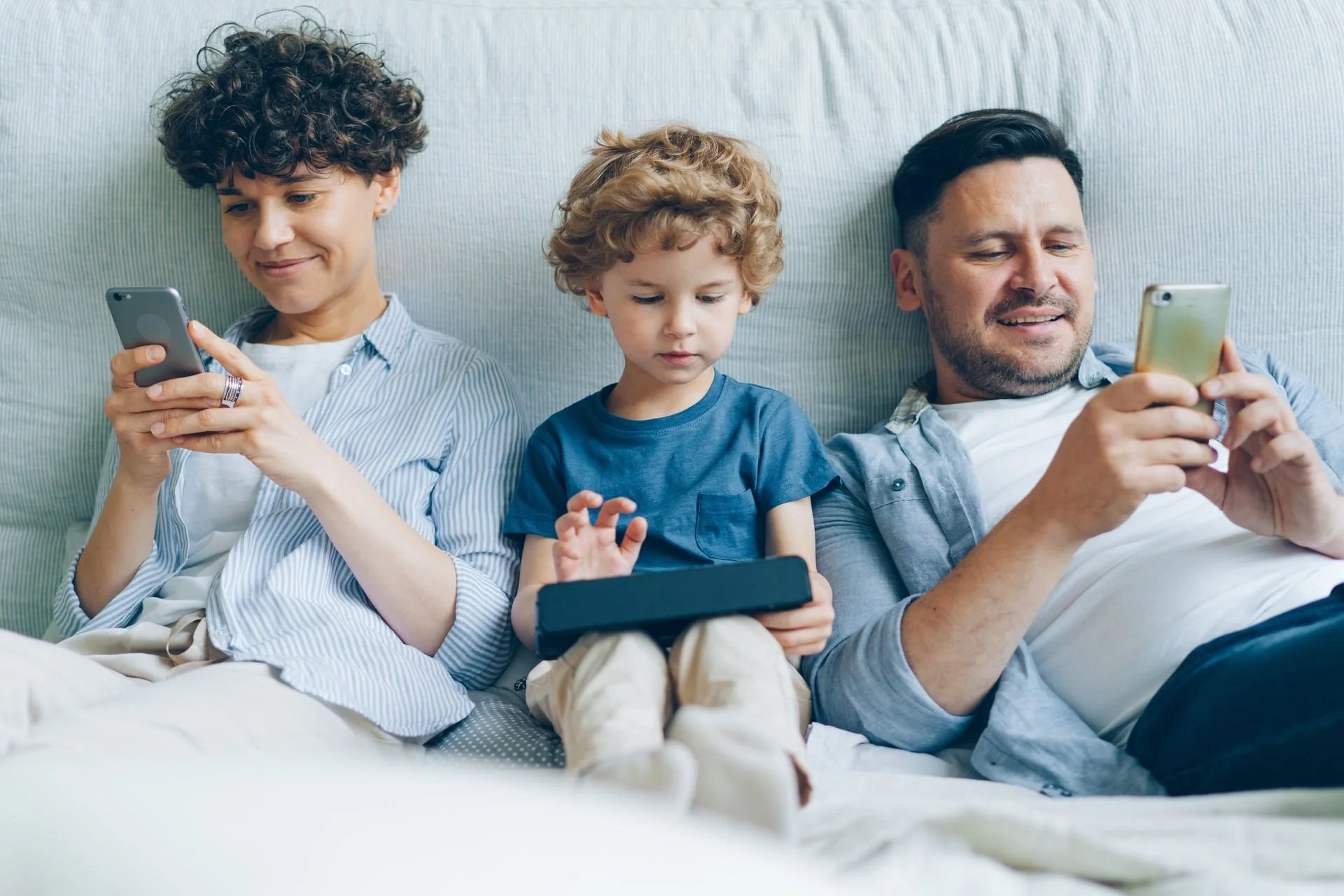 Three people sit next to each other in a bed all being on various electronic devices