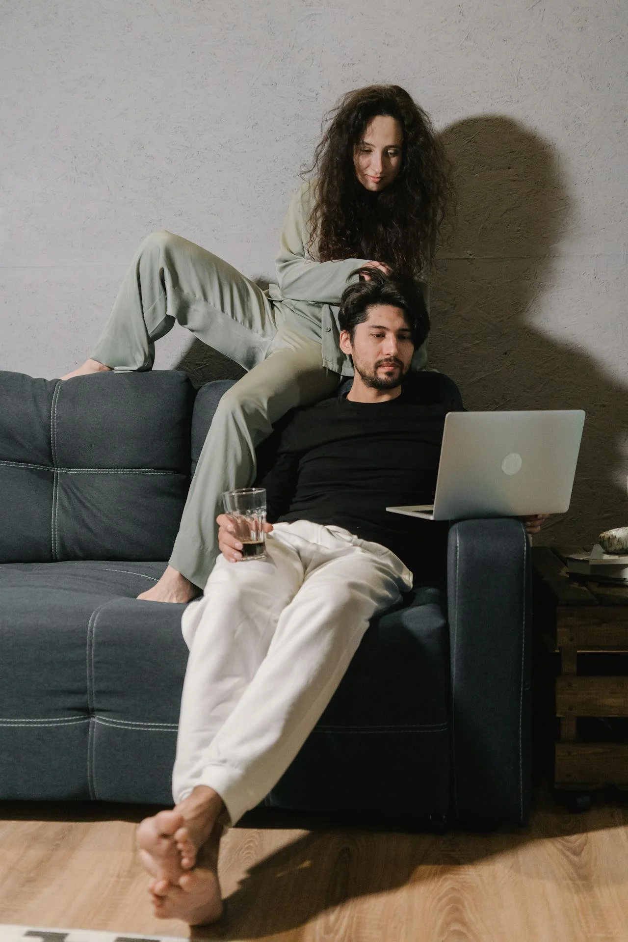 Two people relaxing on a gray couch, one sitting with a laptop and drink, the other leaning behind with a hand on the seated person's head.