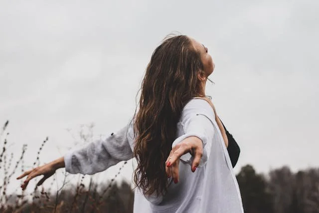 A woman with long hair in a white cardigan stands outdoors with arms outstretched, facing upward, embracing nature on a cloudy day.