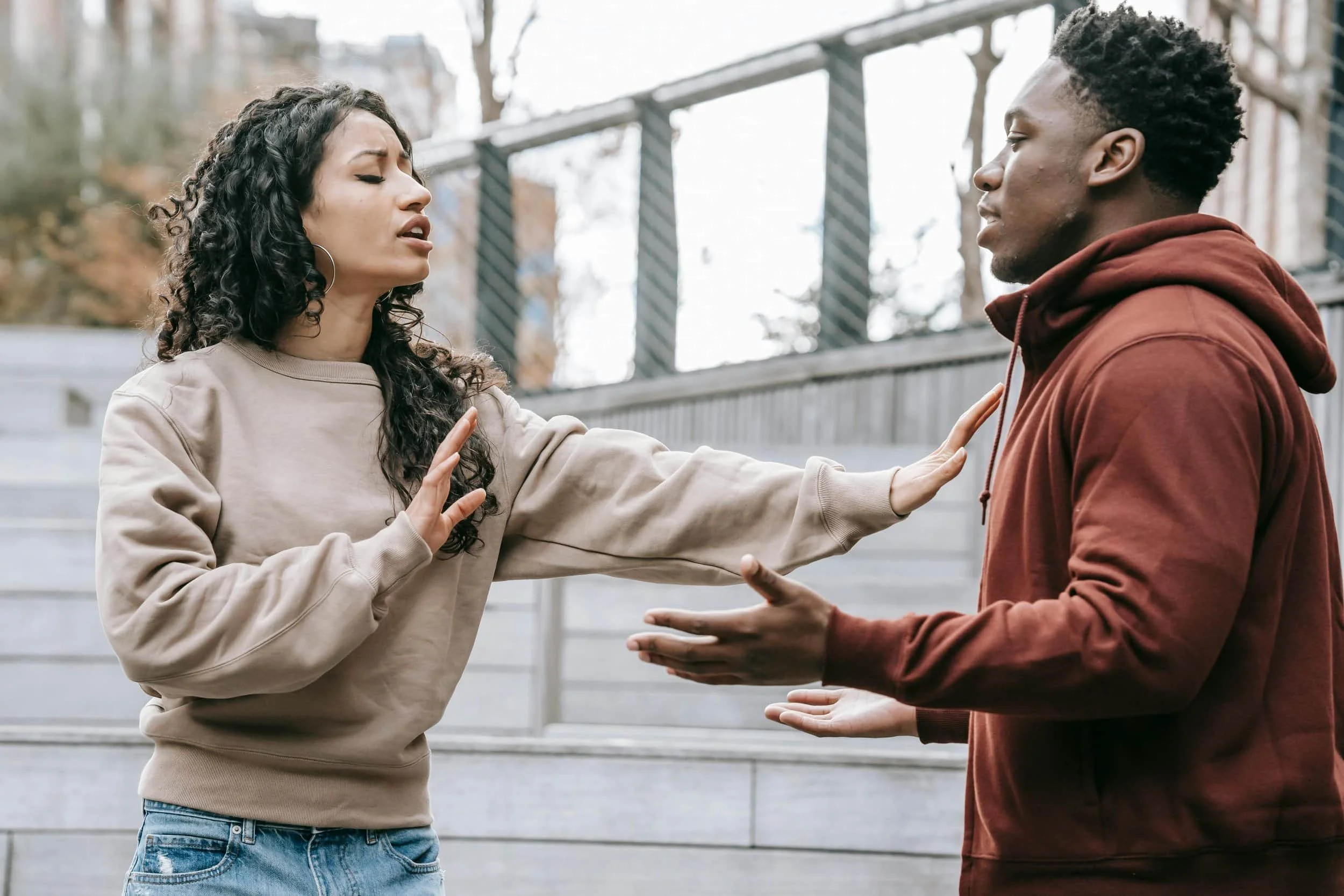 Two people engaged in a serious conversation outdoors, one gesturing with open palms and the other holding up a hand to stop.