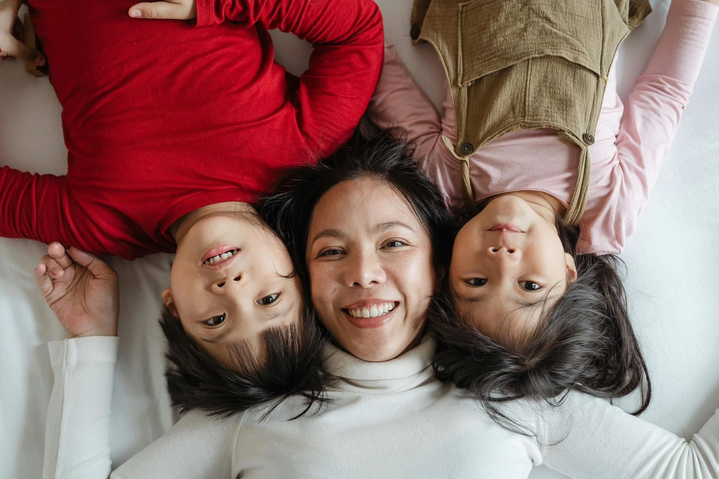 A woman lying on the floor with two children, all smiling and looking up at the camera.
