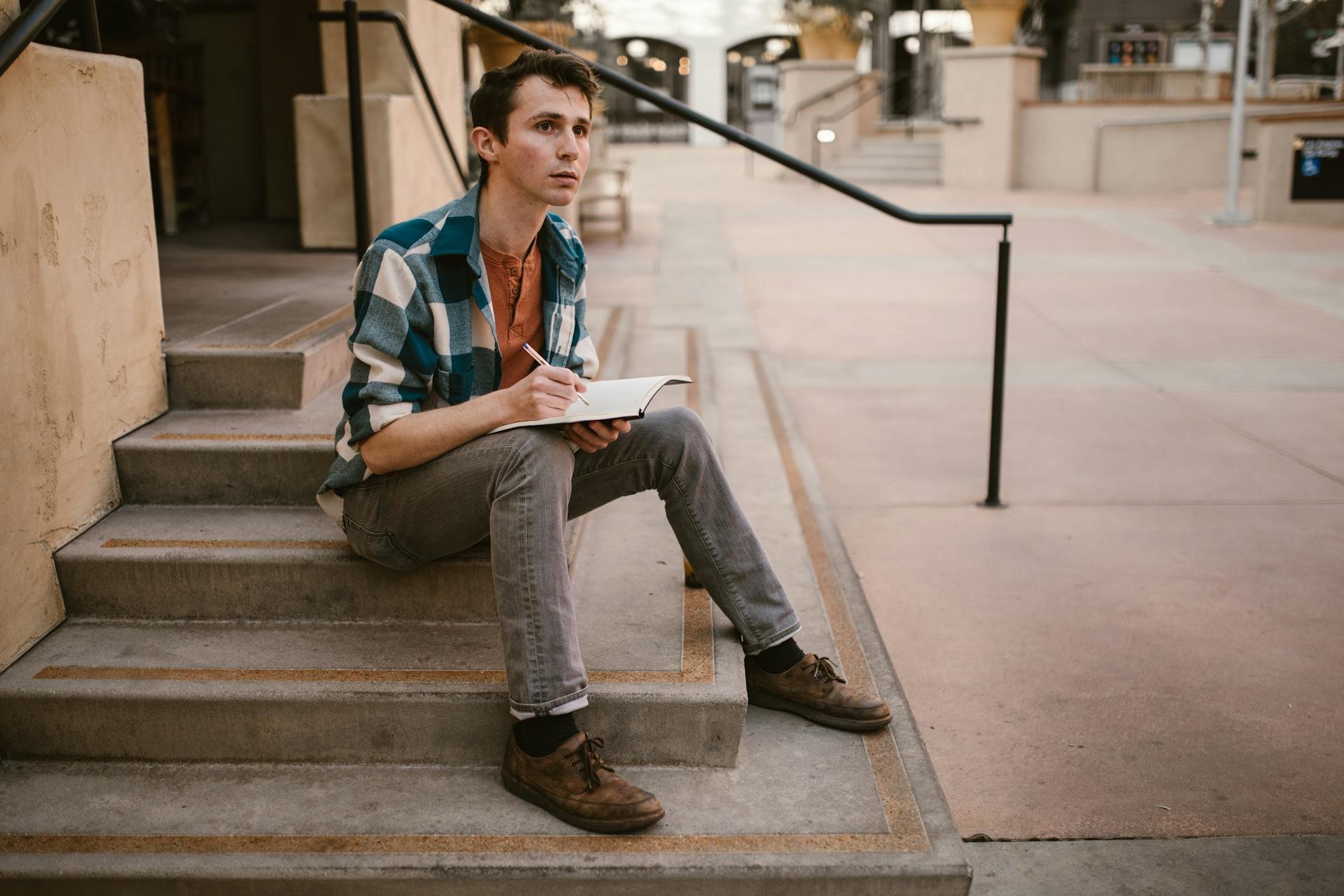 Person in casual clothes sitting on outdoor steps, writing in a notebook, with an empty urban space in the background.