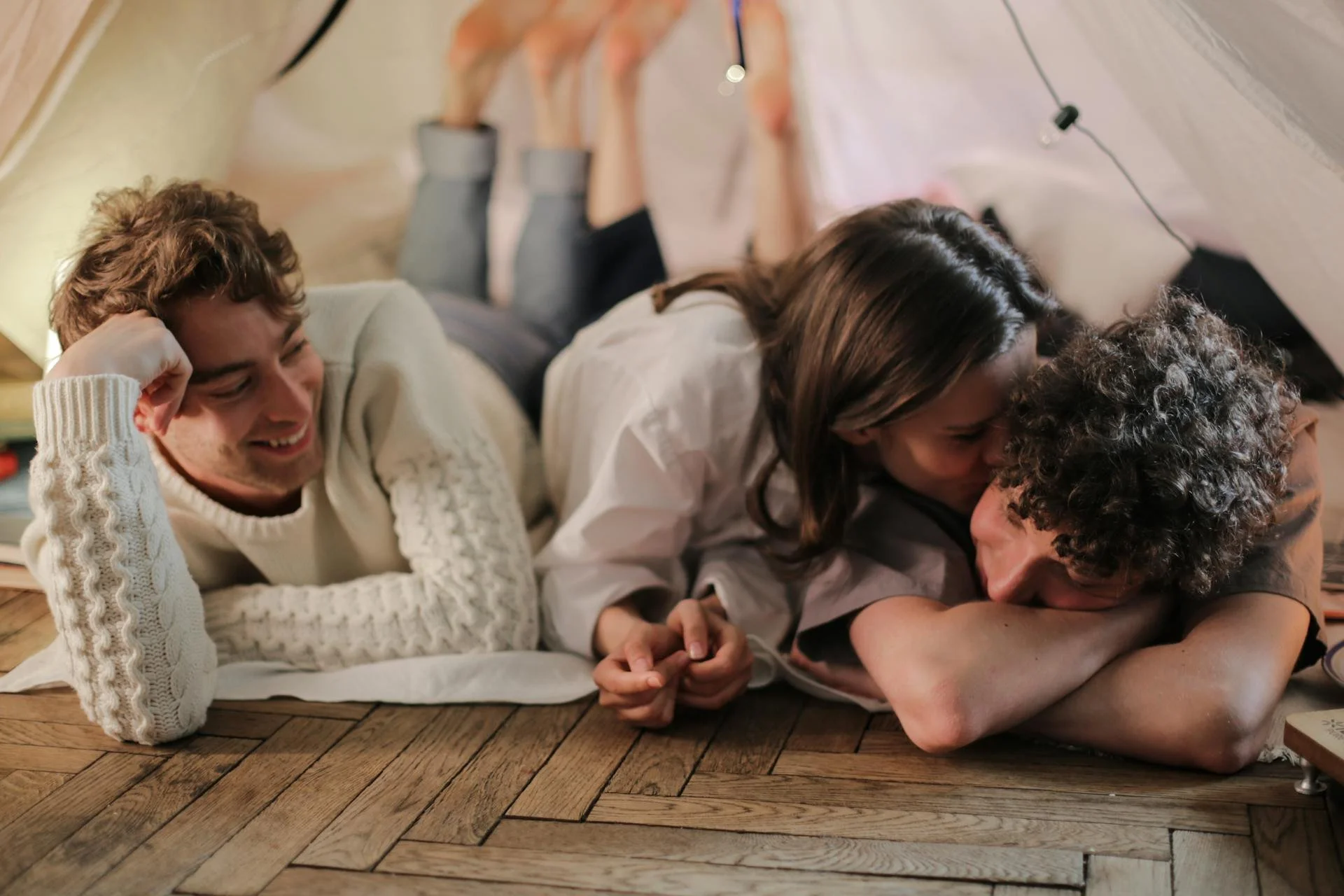 Three friends lying on the floor under a blanket fort, laughing and relaxing together on a wooden floor.