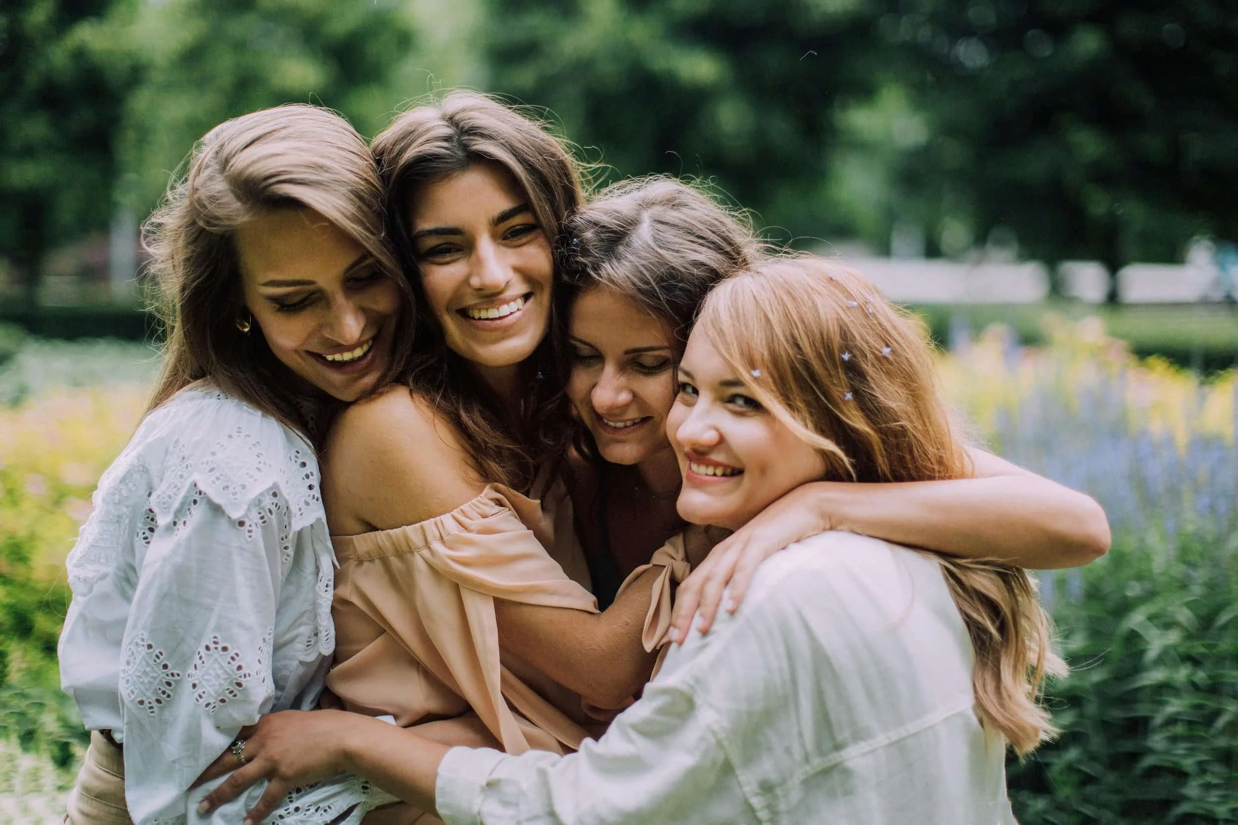 Four women in light-colored clothes hugging outdoors in a lush green garden with flowers.