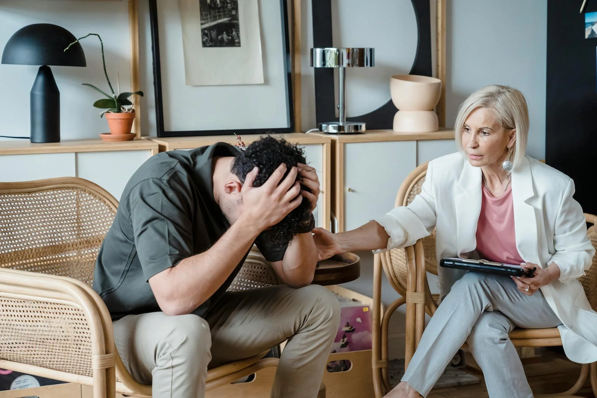 A woman with blonde hair, wearing a light-colored blazer and pants, sits on a chair holding a tablet. A man with curly dark hair, wearing a dark shirt and beige pants, sits on another chair with his head in his hands, visibly upset. The woman is gently holding his arm and appears to be comforting him in a room with modern decor, including artwork, plants, and lamps.