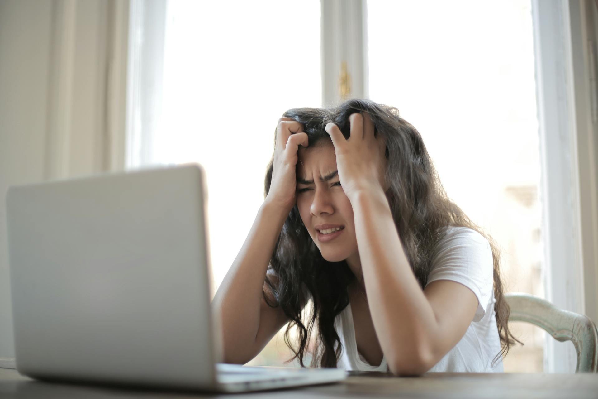 Woman sitting at a desk, holding her head in frustration while looking at a laptop in front of a bright window.