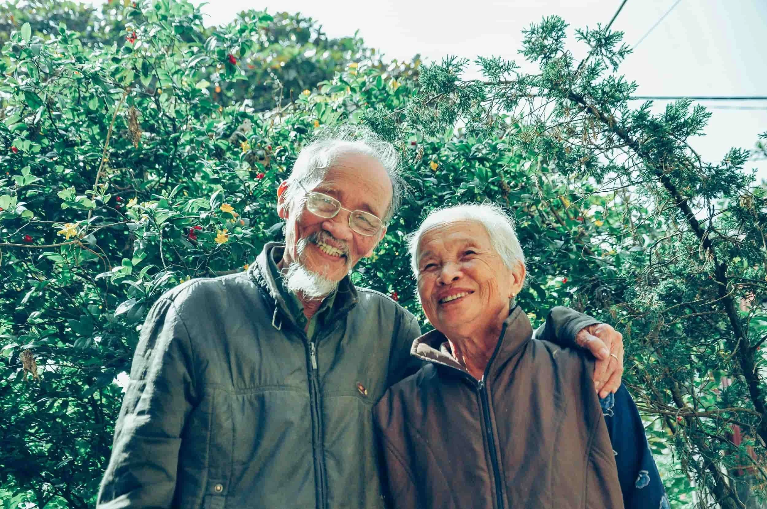 Two elderly people wearing jackets with arms around each other, standing outdoors in front of green foliage.
