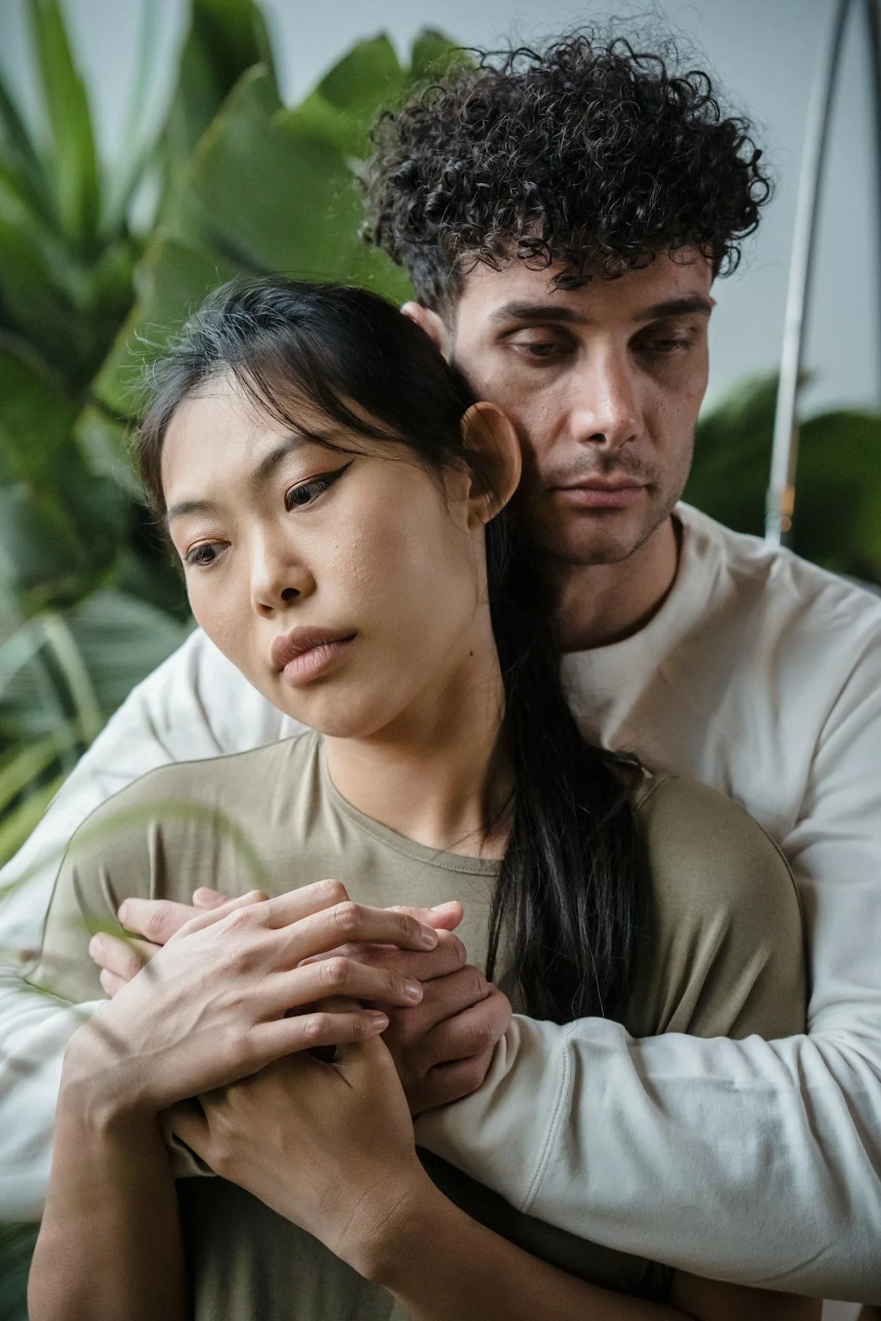 Couple embracing indoors surrounded by green plants, with the person behind wrapping arms around the one in front.