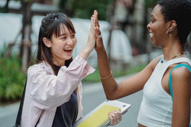 Two people greet each other with high fives on a street, surrounded by greenery and urban scenery. One holds a notebook.