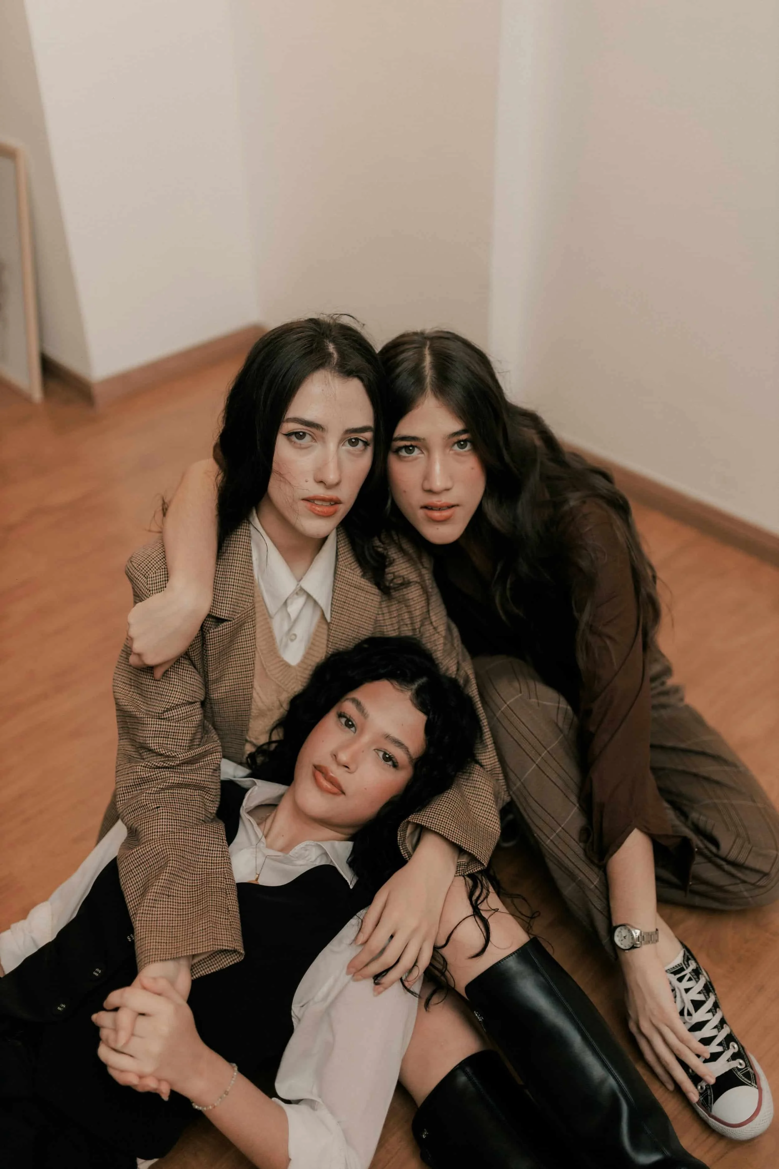 Three women sitting on a wooden floor, closely embracing each other, wearing brown and black outfits with boots and sneakers.