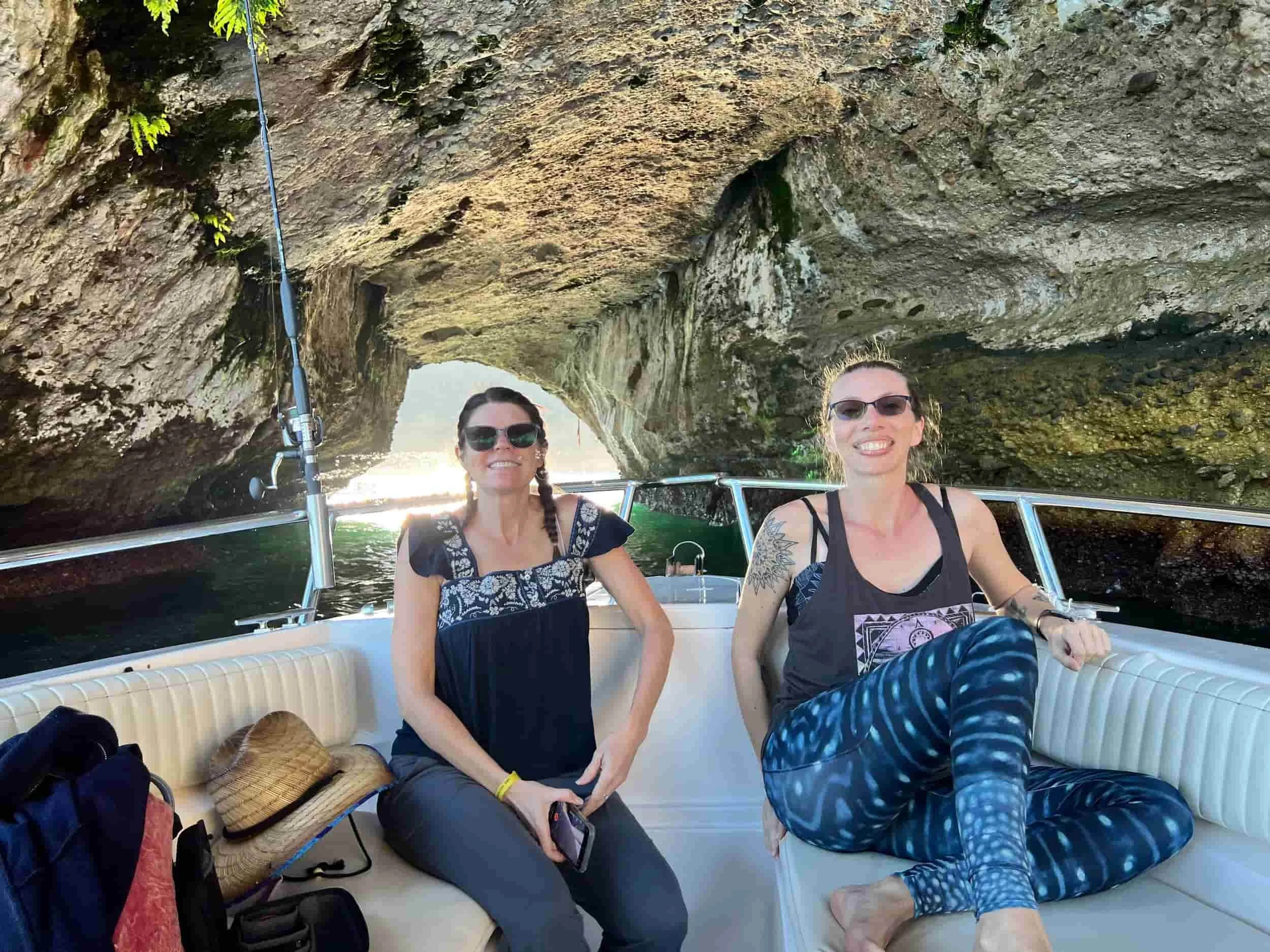 Two people sitting on a boat inside a rocky cave with light streaming through the cave's opening.