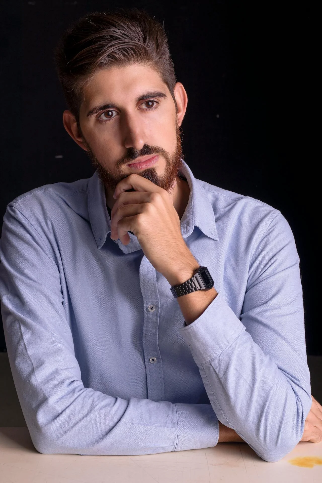 A young man with brown hair and a beard, wearing a light blue shirt and a watch, resting his chin on his hand, looking thoughtfully to the side against a black background.