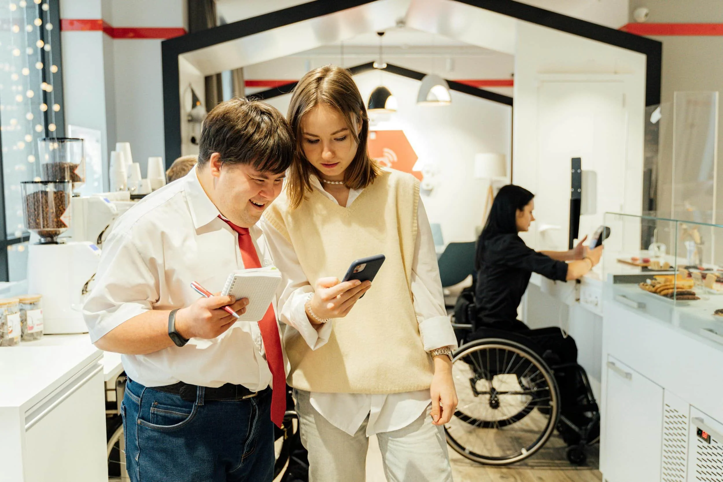 "Two people sharing a happy moment in a café, examining a phone and a notepad, with a wheelchair user visible in the background."
