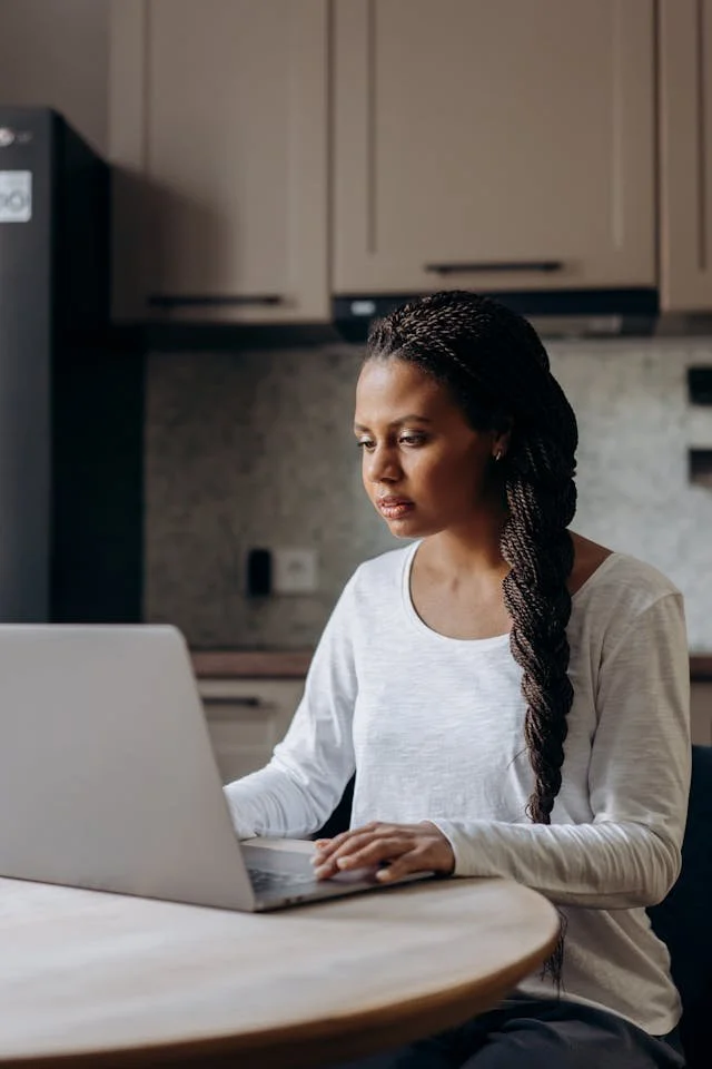 Person with long braided hair working on a laptop at a round kitchen table in a modern kitchen.