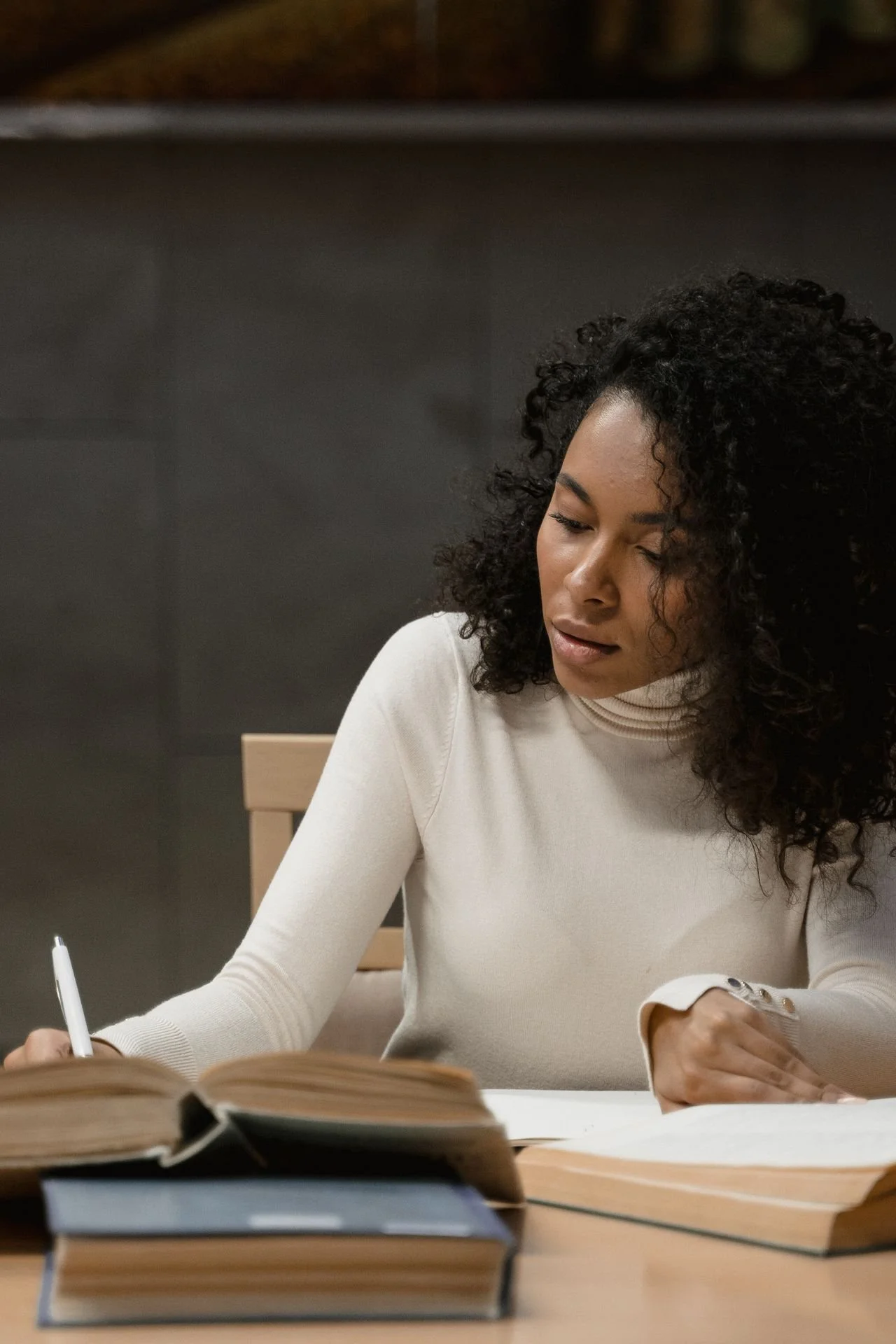 A woman with curly hair writes in a notebook while sitting at a table with open books.