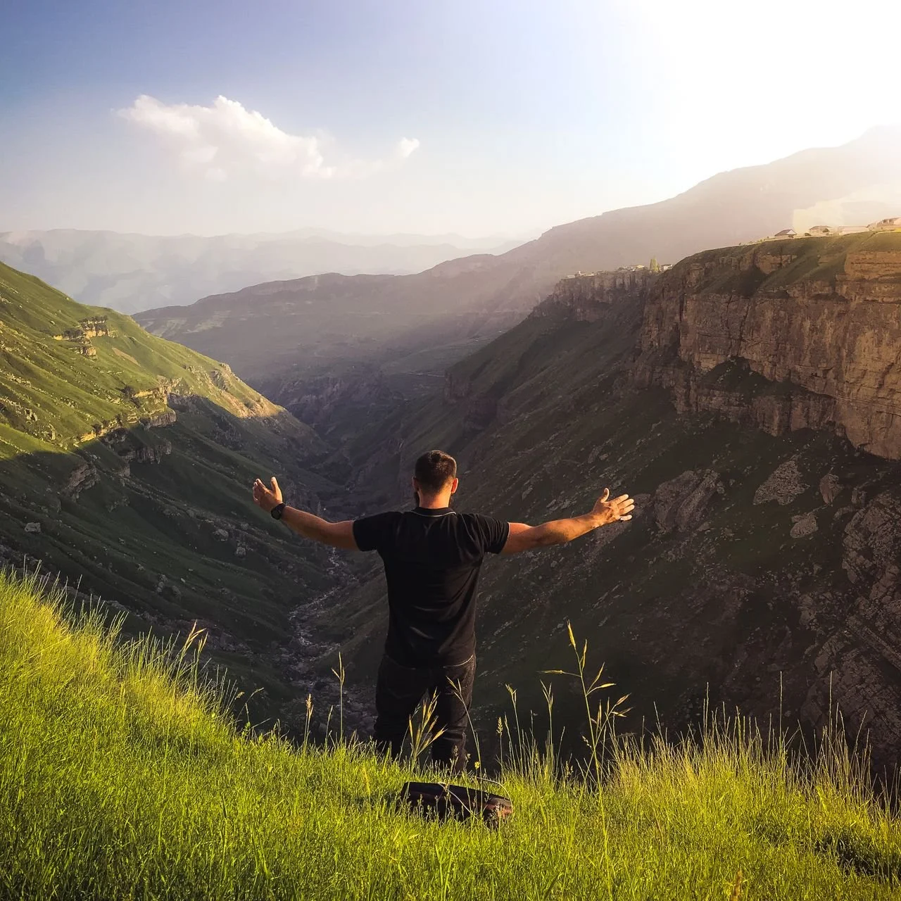 A person stands on green grass with arms wide, overlooking a vast mountain valley in sunlight.