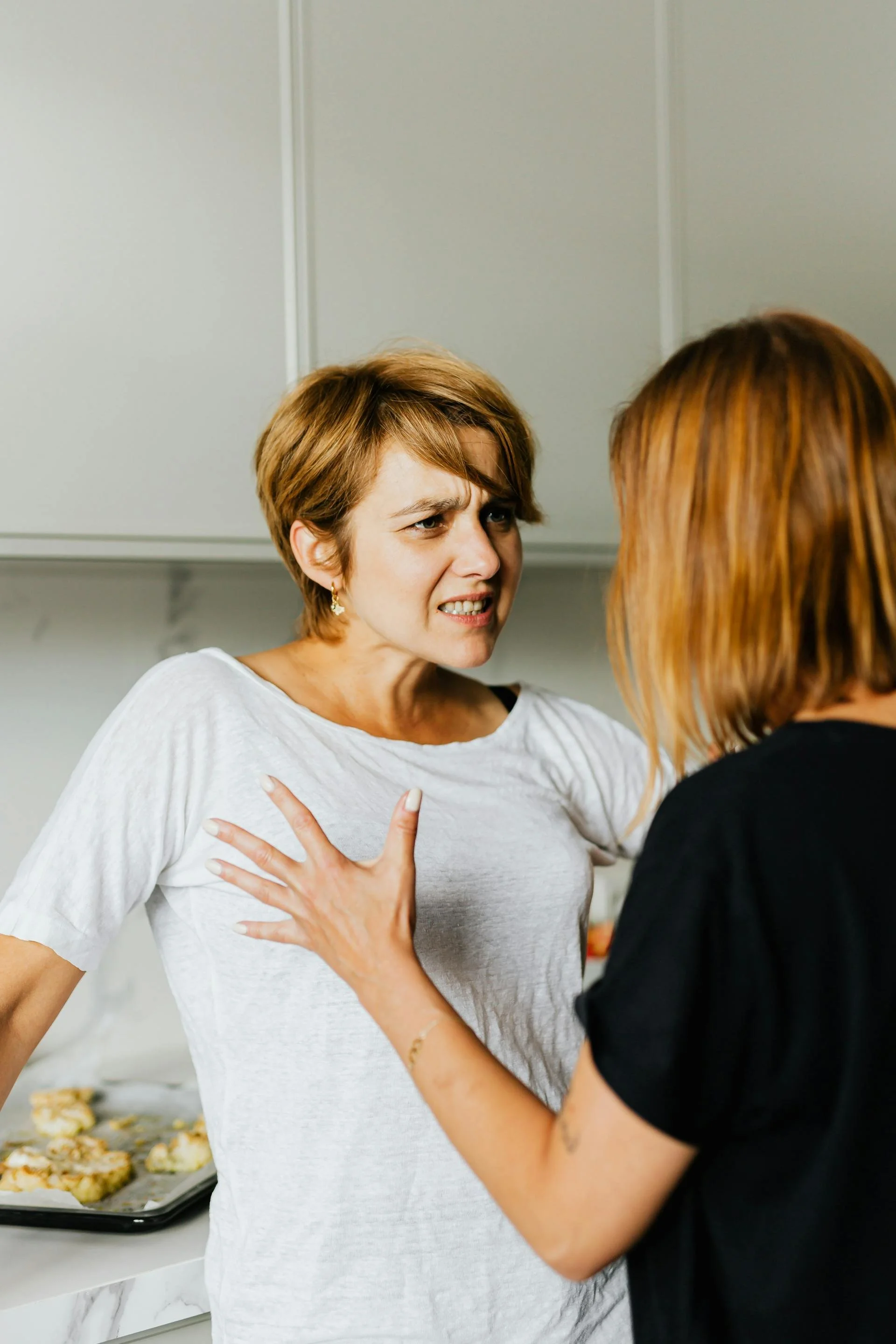Two women in a kitchen, one with hand on the other's chest, engaged in an intense conversation.