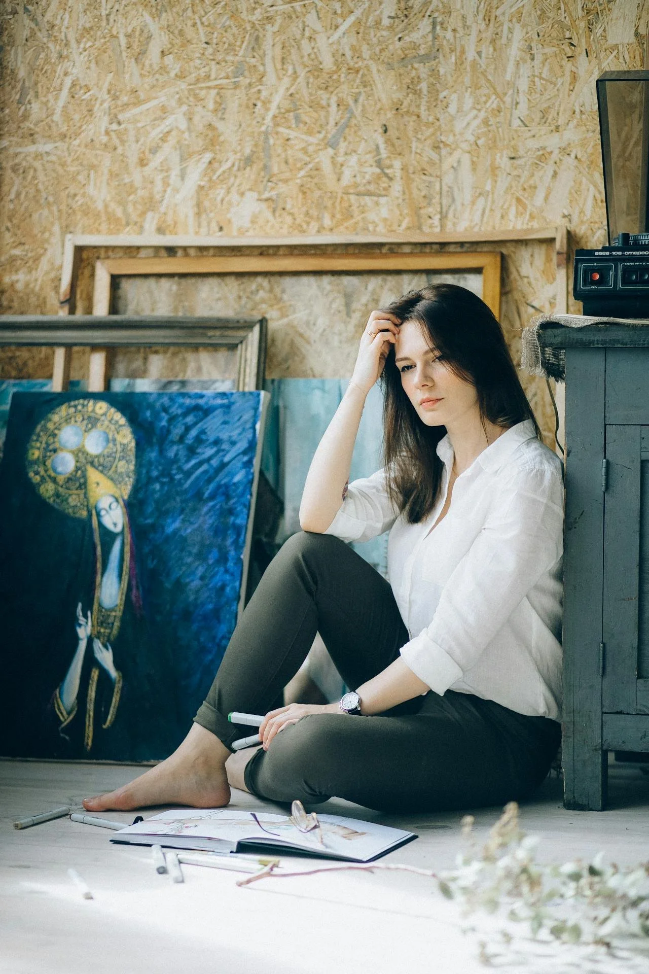 Woman sitting on the floor barefoot, surrounded by sketches and paintings, in a cozy art studio with wooden walls.