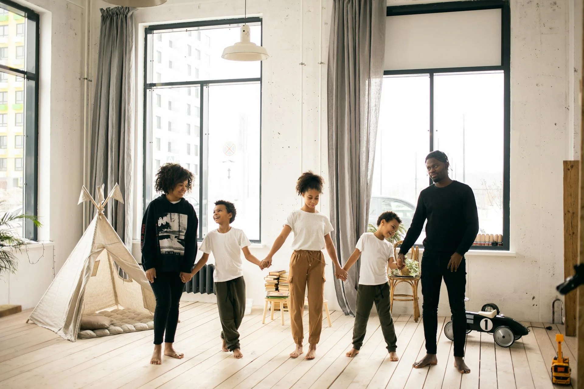 A family of five holding hands and standing barefoot in a bright, spacious room with large windows and a play tent.