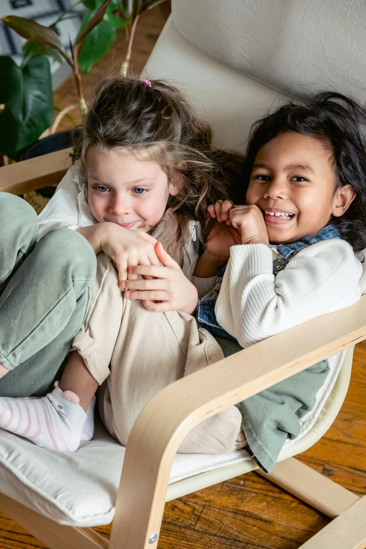 Two young children sitting on a beige cushioned wooden rocking chair, smiling and relaxing indoors with wooden flooring and green plants in the background.