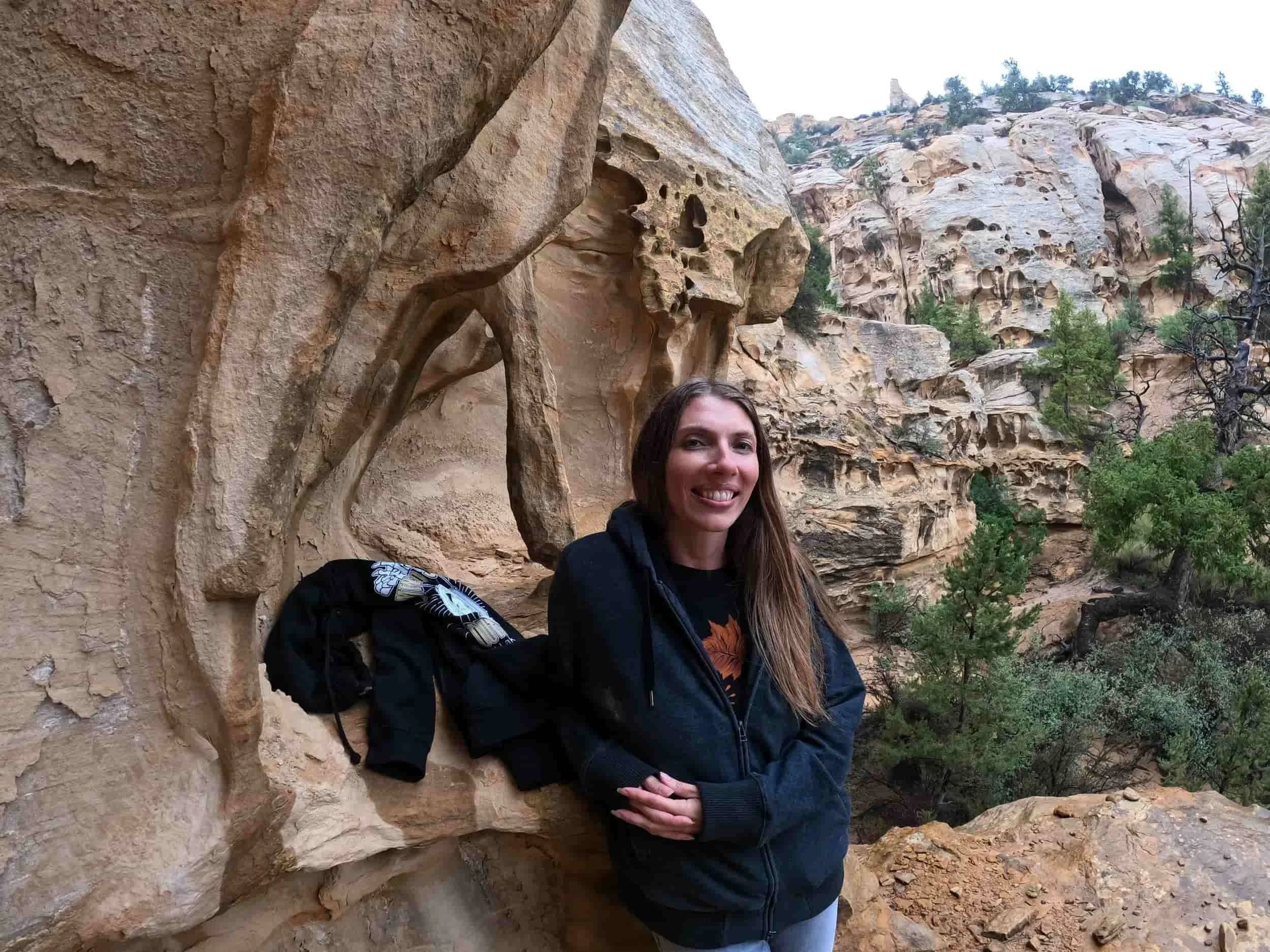 Person in dark hoodie standing against eroded sandstone rock formation with trees and cliffs in the background.