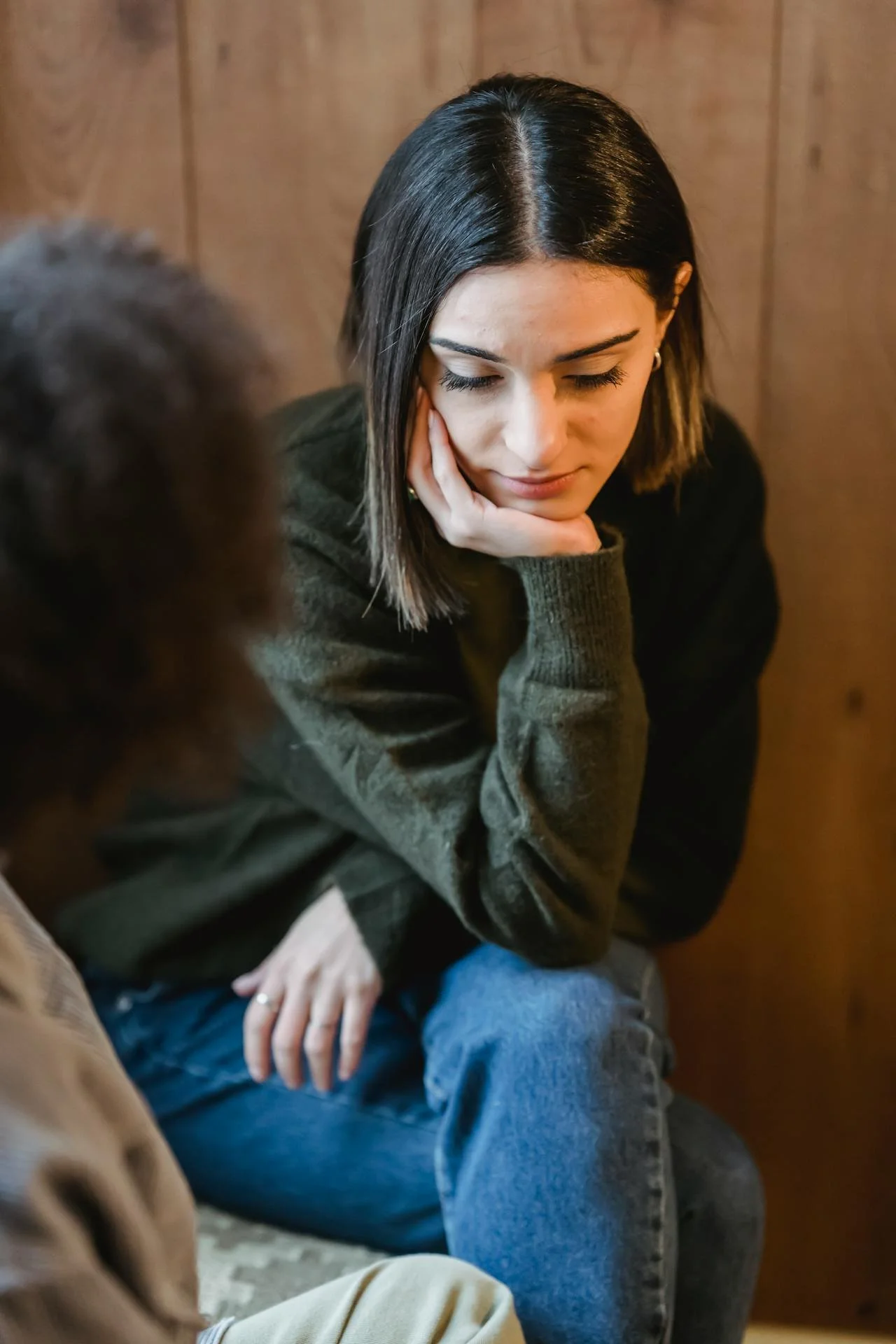 A person in a dark sweater sits in a chair with her chin resting on her hand in deep thought and contemplation
