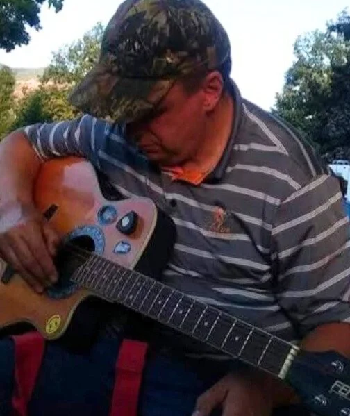 A man in a camo hat plays an acoustic guitar outdoors, surrounded by trees, capturing a moment of music and nature.