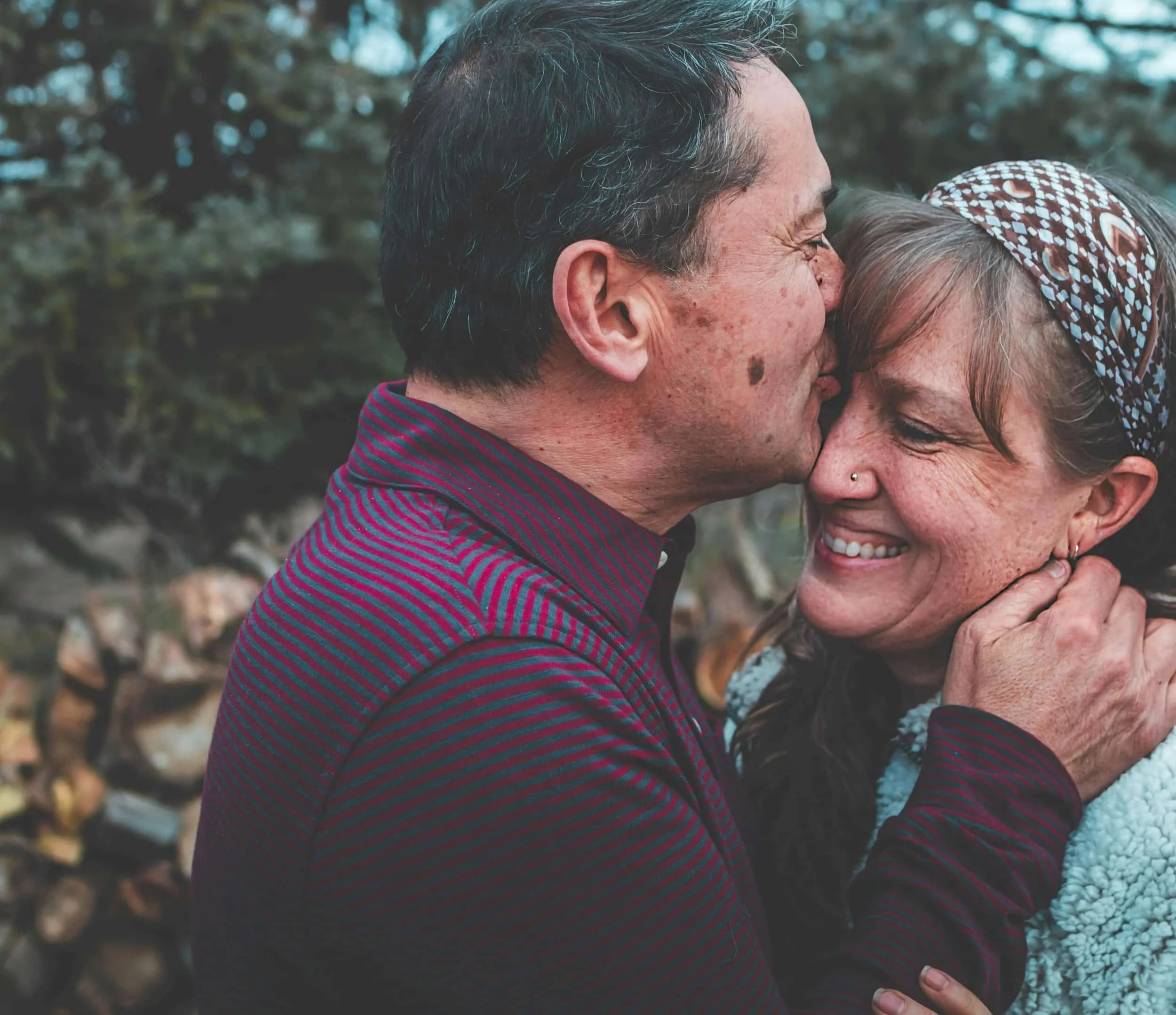 Man in a striped shirt gently holding and kissing woman's face outdoors with blurred green foliage and woodpile in background.