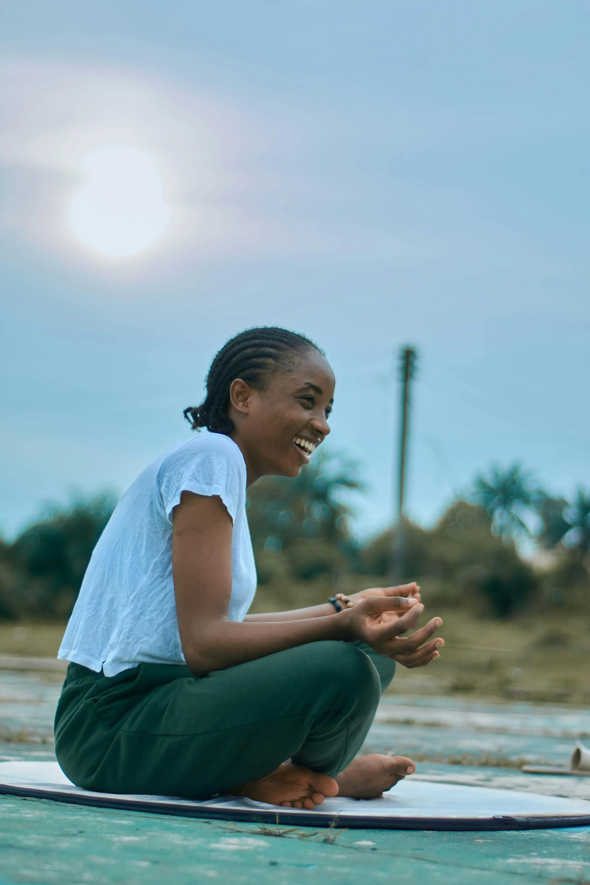 A woman with cornrow braids and a white shirt smiling and practicing yoga outdoors on a mat near water with trees and a utility pole in the background.