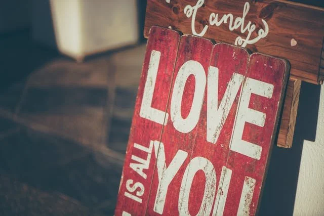 A rustic red wooden sign displaying the phrase "Love is all you need," with decorative accents and a candy shop context.