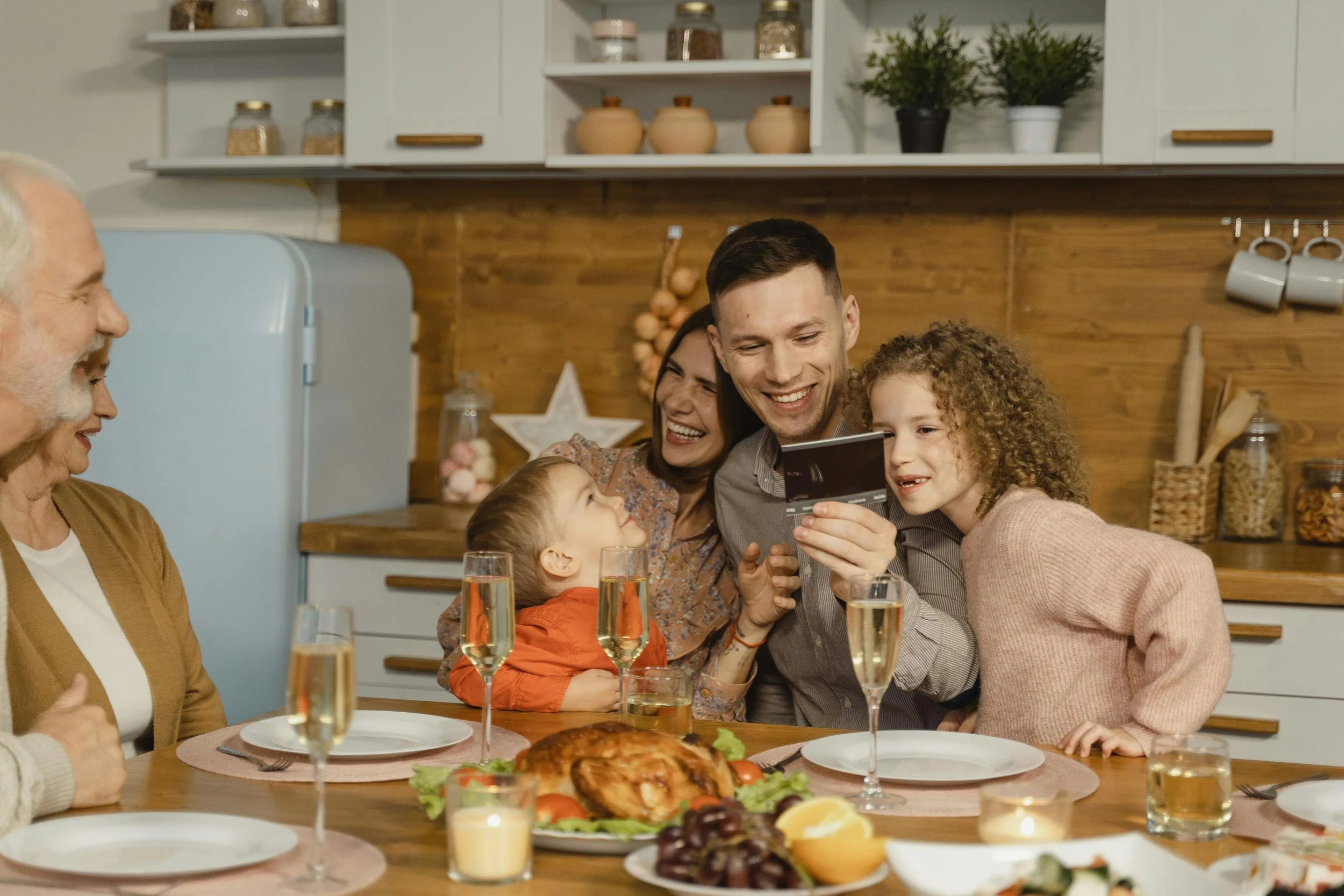 A family gathers around a dining table filled with food, toasting with glasses of sparkling drinks, and sharing a moment of joy.