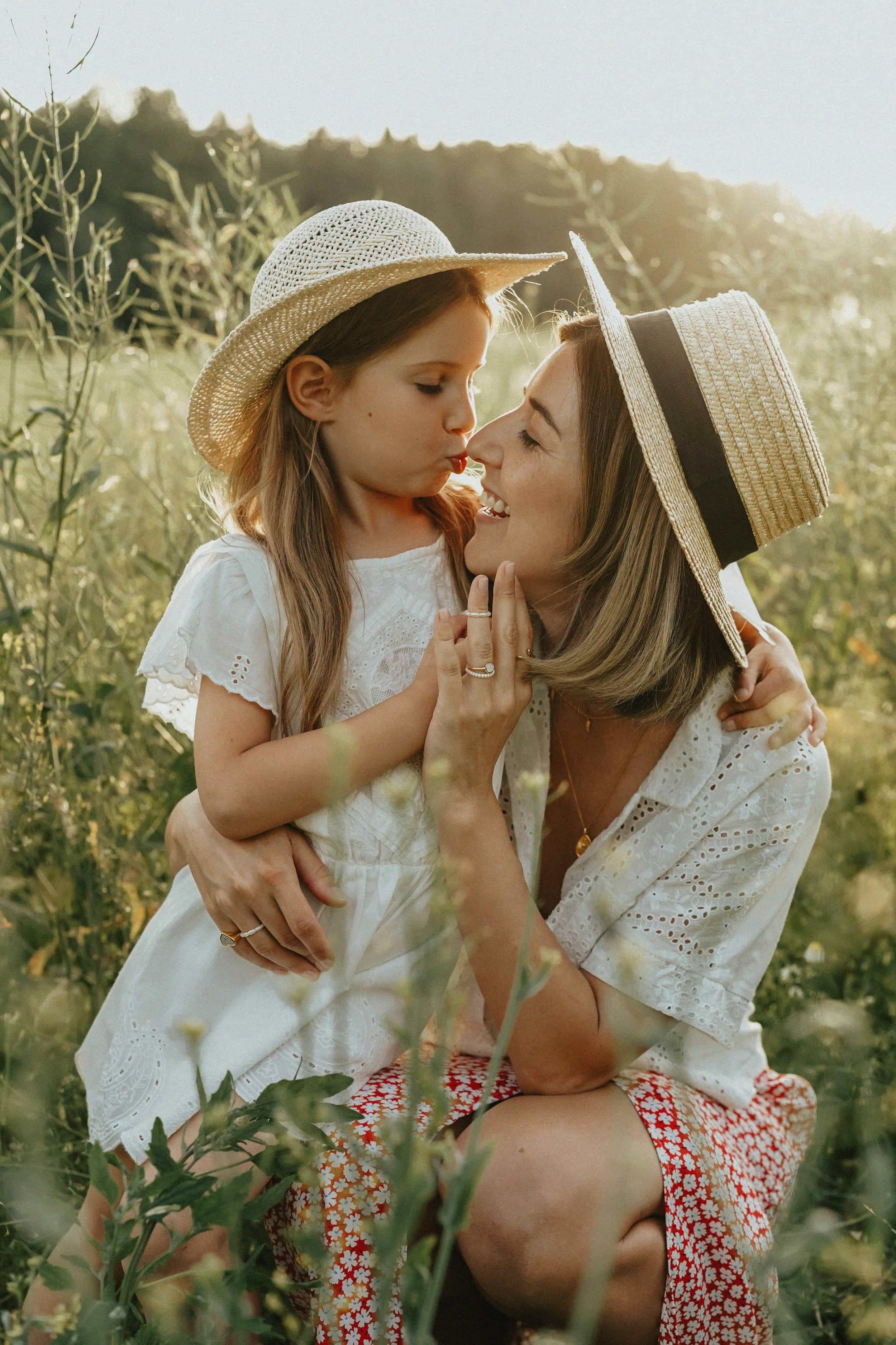 A woman and a young girl, possibly mother and daughter, wearing summer hats and white clothing, sharing a moment together outdoors in a natural setting with green foliage and sunlight.
