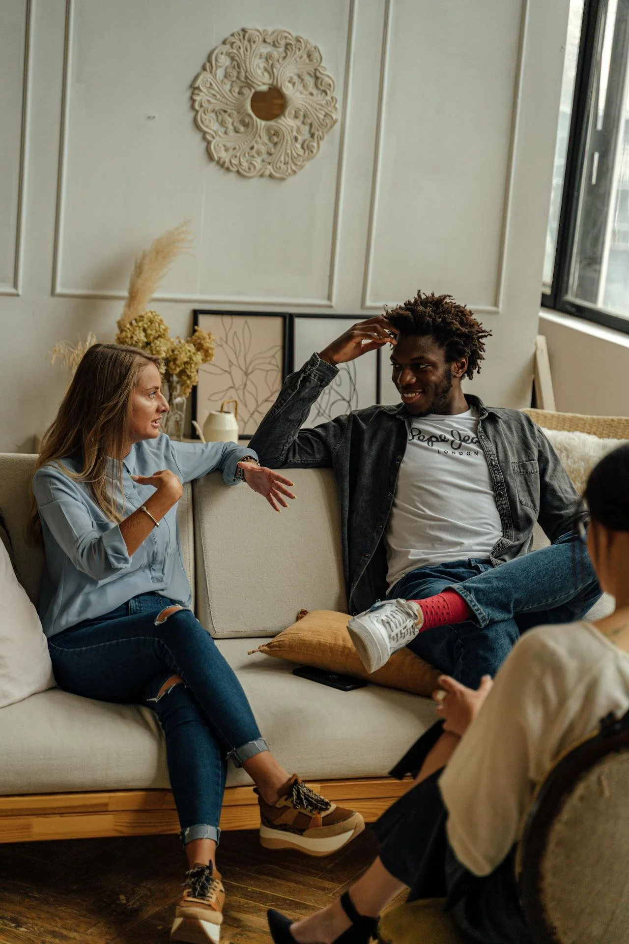 Three people in a casual living room setting engaged in conversation, seated on a couch and chair near a window.